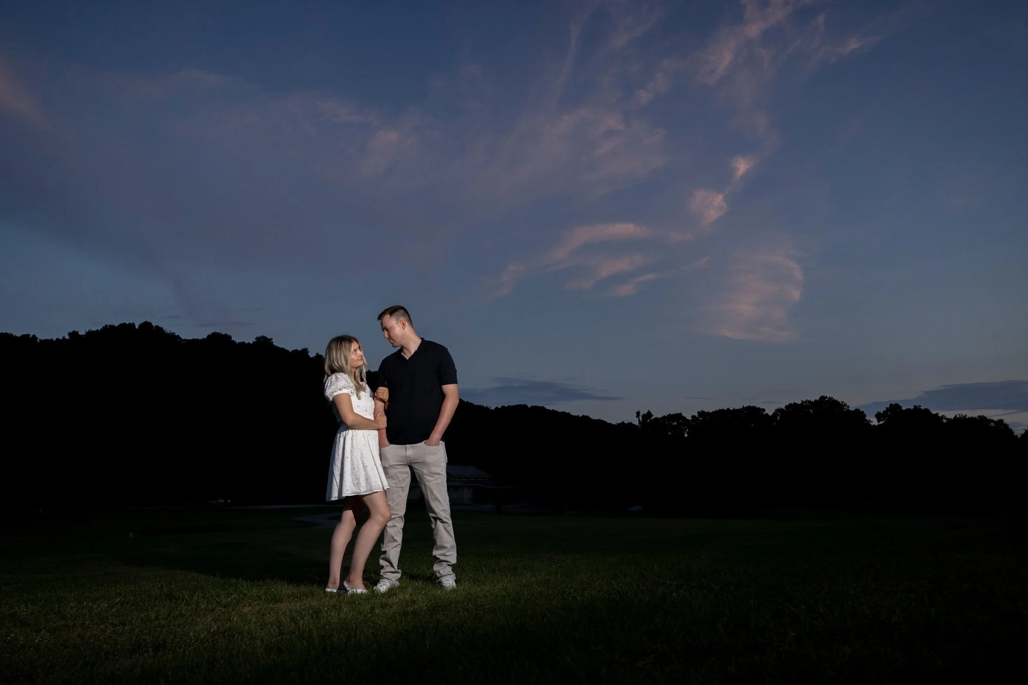 A young woman and man standing close together outdoors during dusk, holding hands and looking at each other, with a dark silhouetted landscape and a partly cloudy sky in the background.
