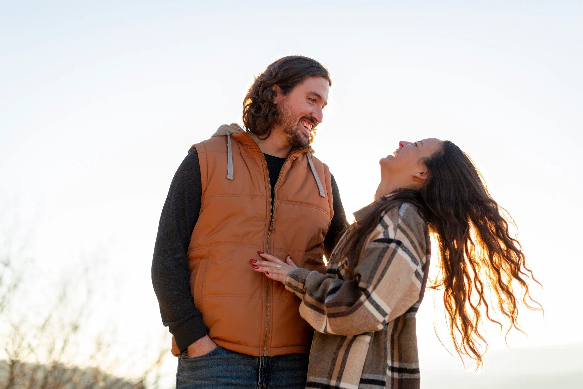 A man and woman smiling and looking at each other outdoors during sunset, standing close with the woman's hand on the man's chest. The man has long curly hair and is wearing a brown vest over a black shirt, while the woman has long wavy hair and is w