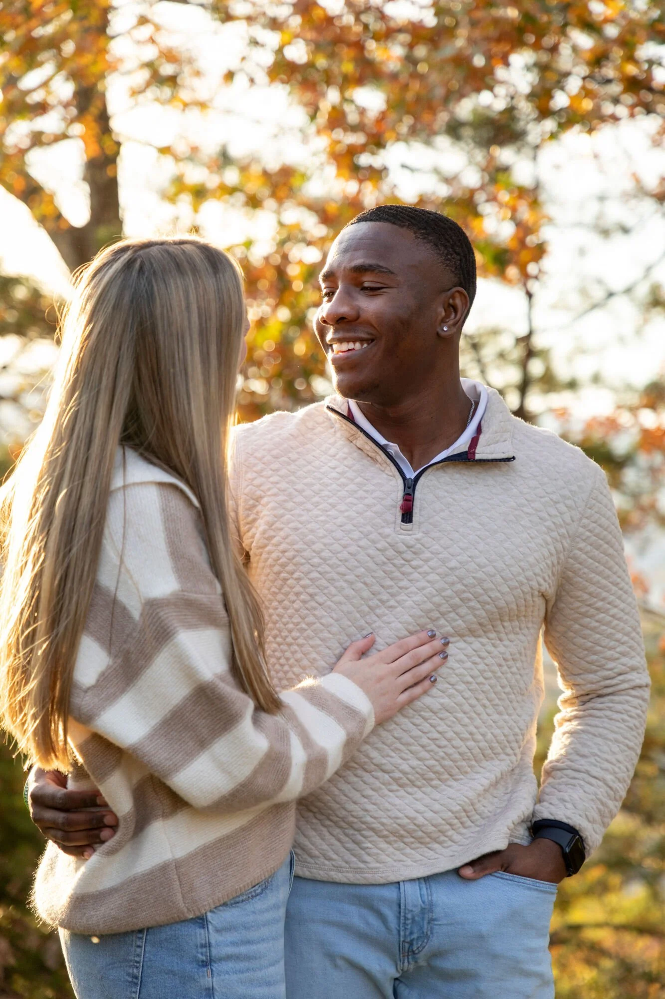A couple standing outdoors during autumn, with the woman touching the man's chest and both smiling at each other, background of fall foliage.