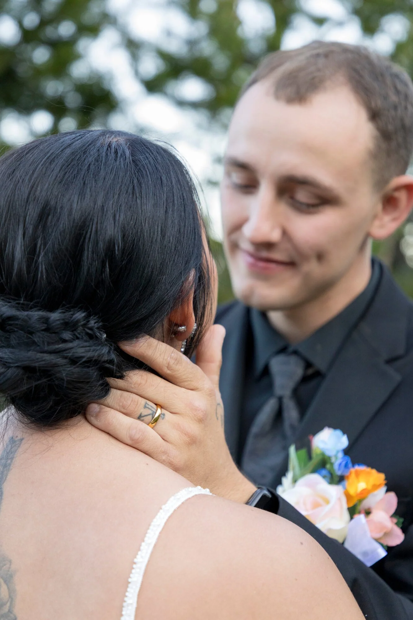 A bride and groom sharing a kiss outdoors, with the bride touching the groom's face and holding a bouquet of flowers.