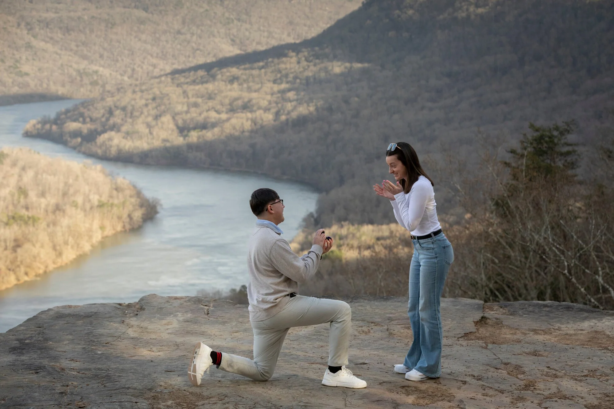 A man on one knee proposing to a woman outdoors near a river with hills in the background. The woman is smiling and clapping her hands, while the man is holding a small box, possibly with a ring, on one knee.