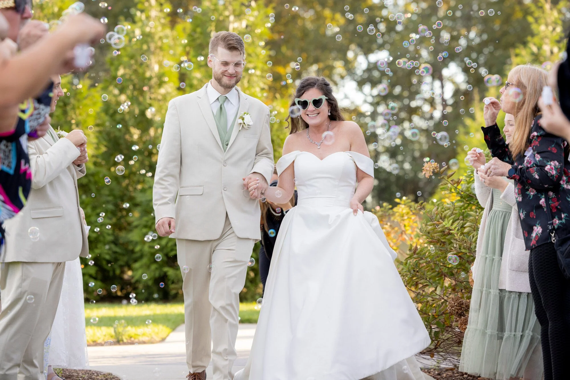 A bride and groom walking outdoors during a celebration, surrounded by friends and family, with bubbles floating around them, greenery in the background, and sunlight filtering through the leaves.