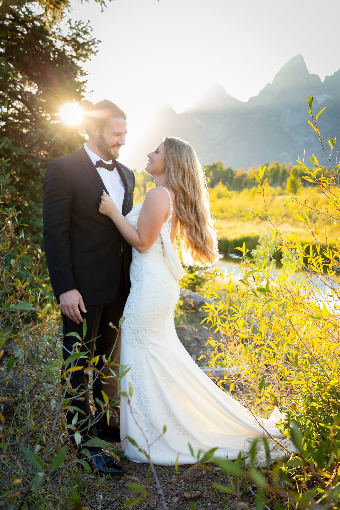 A bride and groom standing outdoors in a natural setting during sunset, smiling at each other, with mountains in the background.