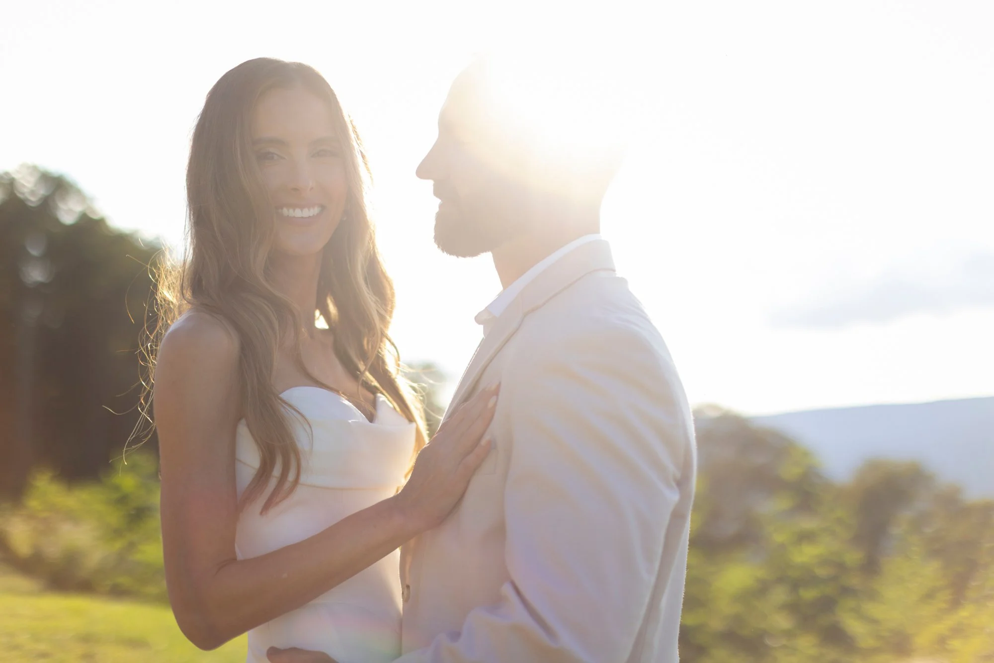 A smiling woman with long wavy hair and a man in a white suit outdoors during sunset, with sunlight creating a bright glow.