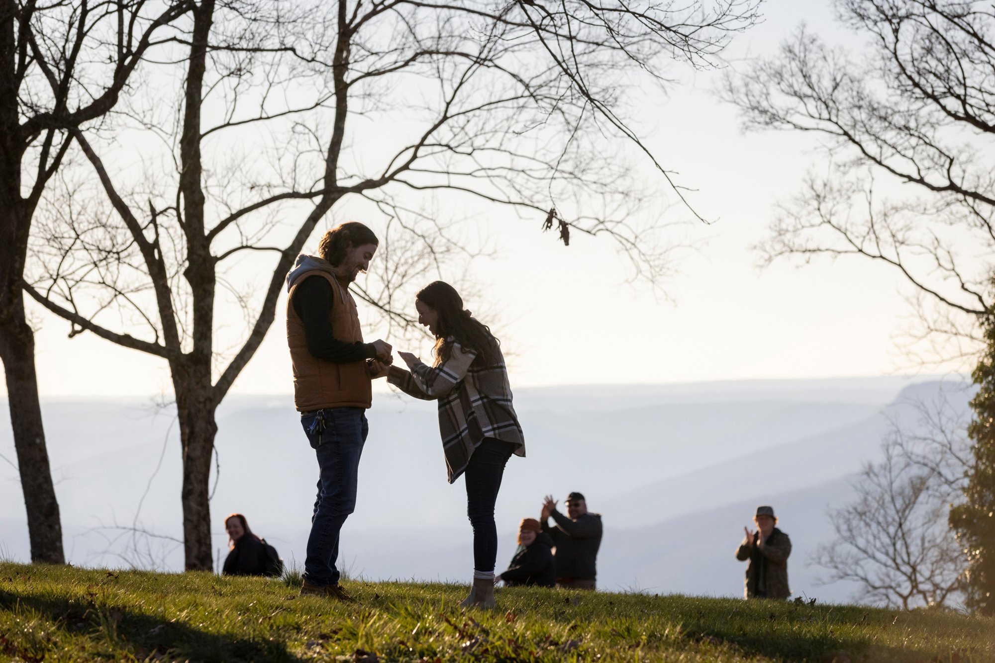 A group of people outdoors in a park on a clear day, with one man and woman standing on the grass exchanging a small item or gift, surrounded by leafless trees and other people in the background.