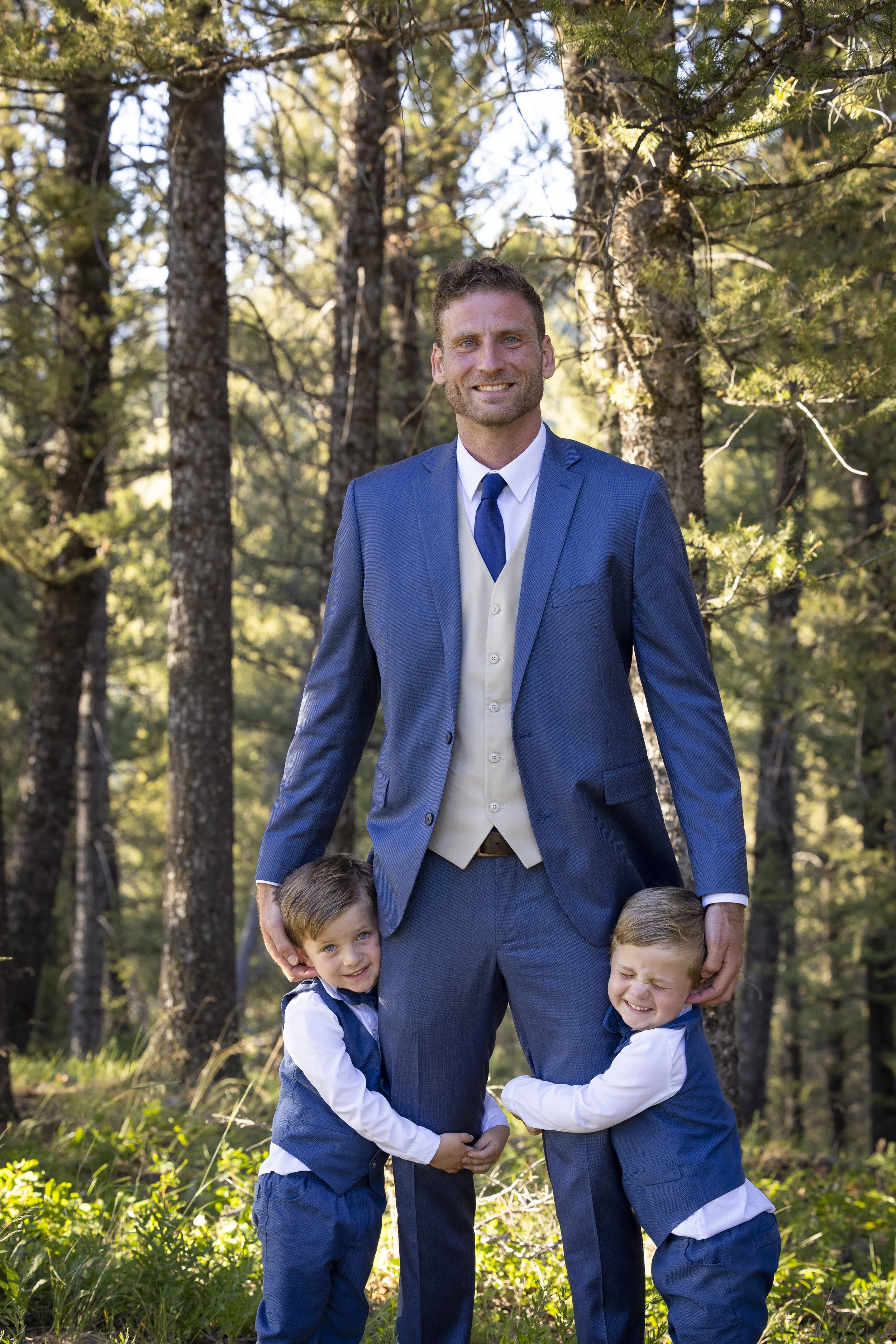 A man in a blue suit with two young boys in matching blue vests and white shirts standing outdoors among trees in a forested area.