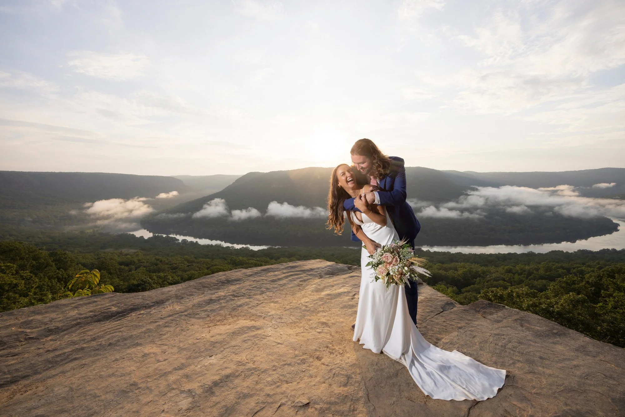 A newlywed couple is embracing on a large rock with a scenic mountain landscape and a cloudy sky in the background. The woman is wearing a white wedding dress and holding a bouquet, and the man is in a dark suit. They are smiling and appear joyful.