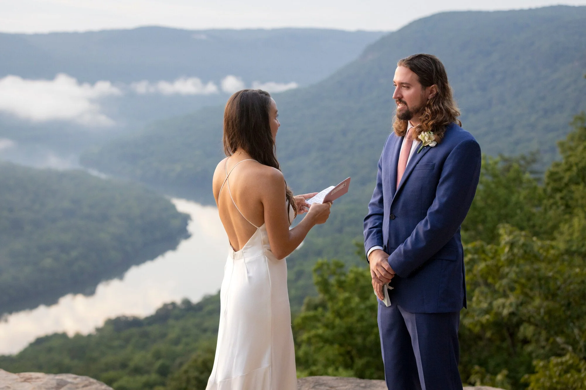 A woman in a white dress is saying her wedding vows to a man in a blue suit during an outdoor wedding ceremony with mountains in the background.