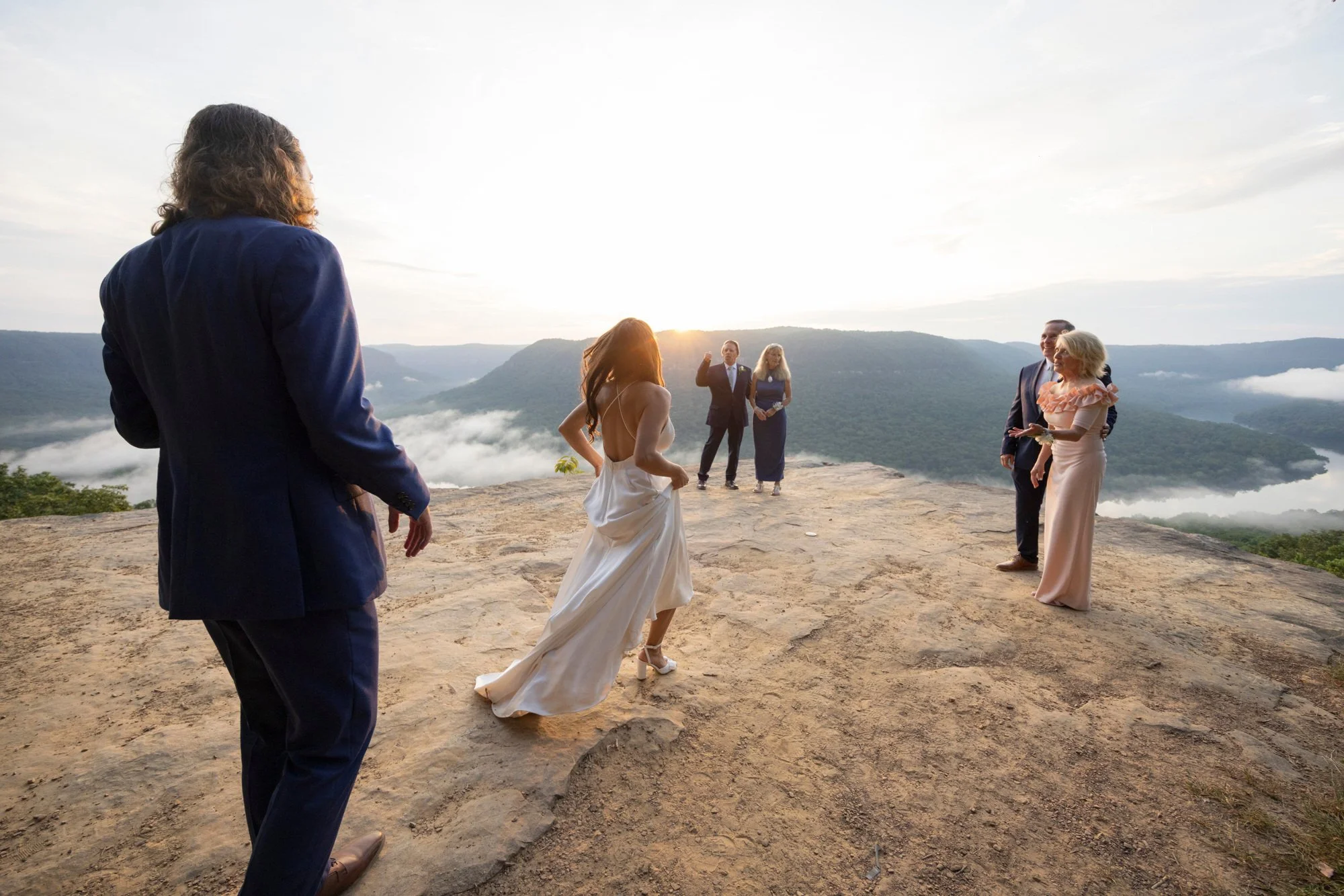 A wedding ceremony taking place outdoors on a mountain ridge at sunrise, with a bride dancing in her wedding dress and guests dressed formally watching.