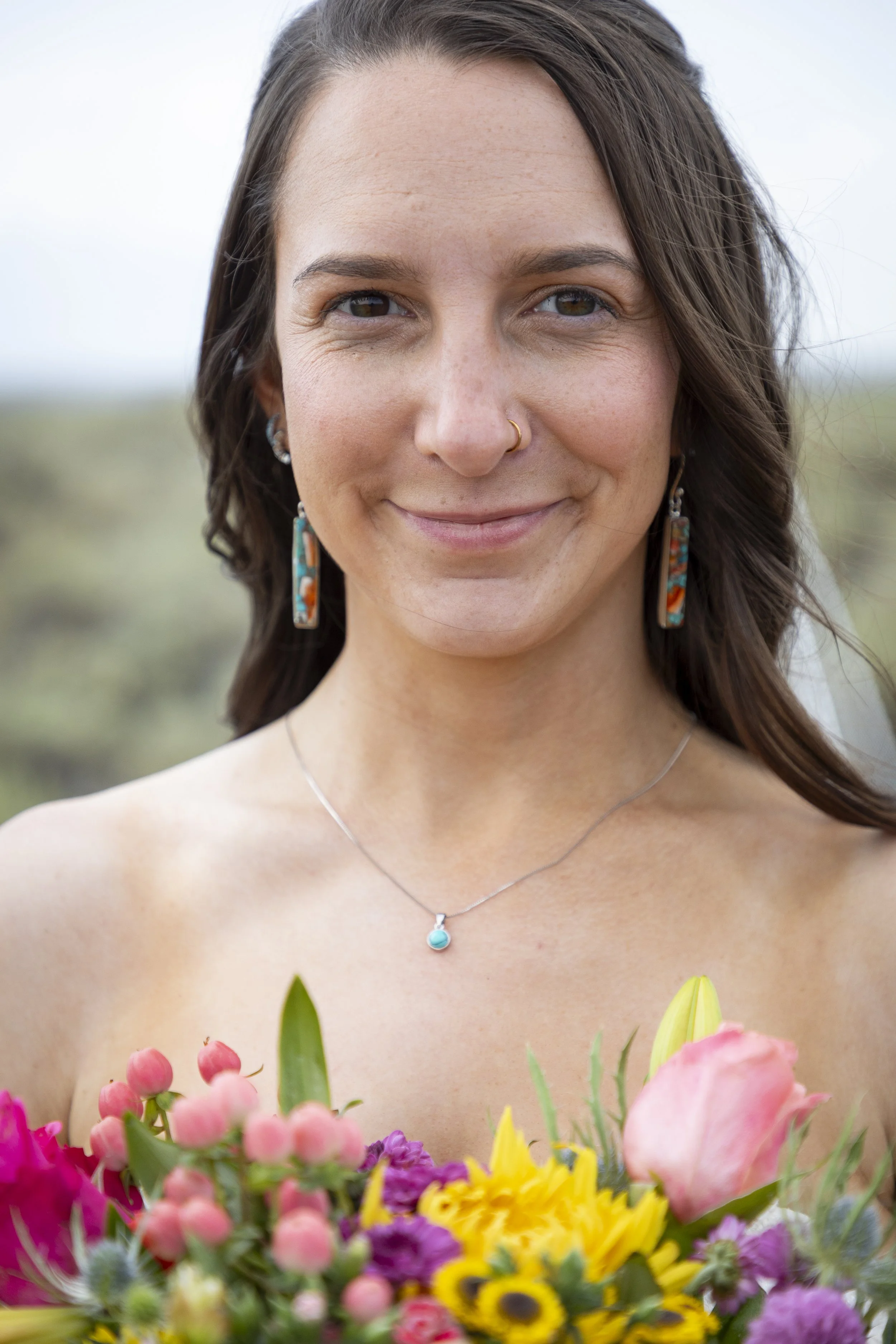 A woman with brown hair, wearing earrings and a necklace, smiling while holding a bouquet of colorful flowers outdoors.
