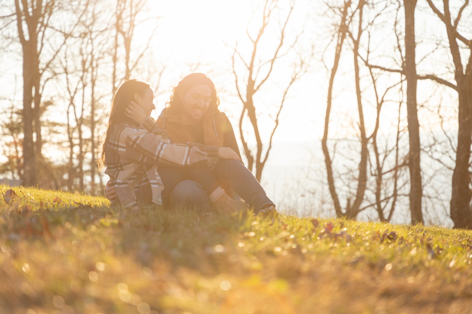 A man and a woman sitting on the grass in a wooded area during sunset, sharing a joyful moment.