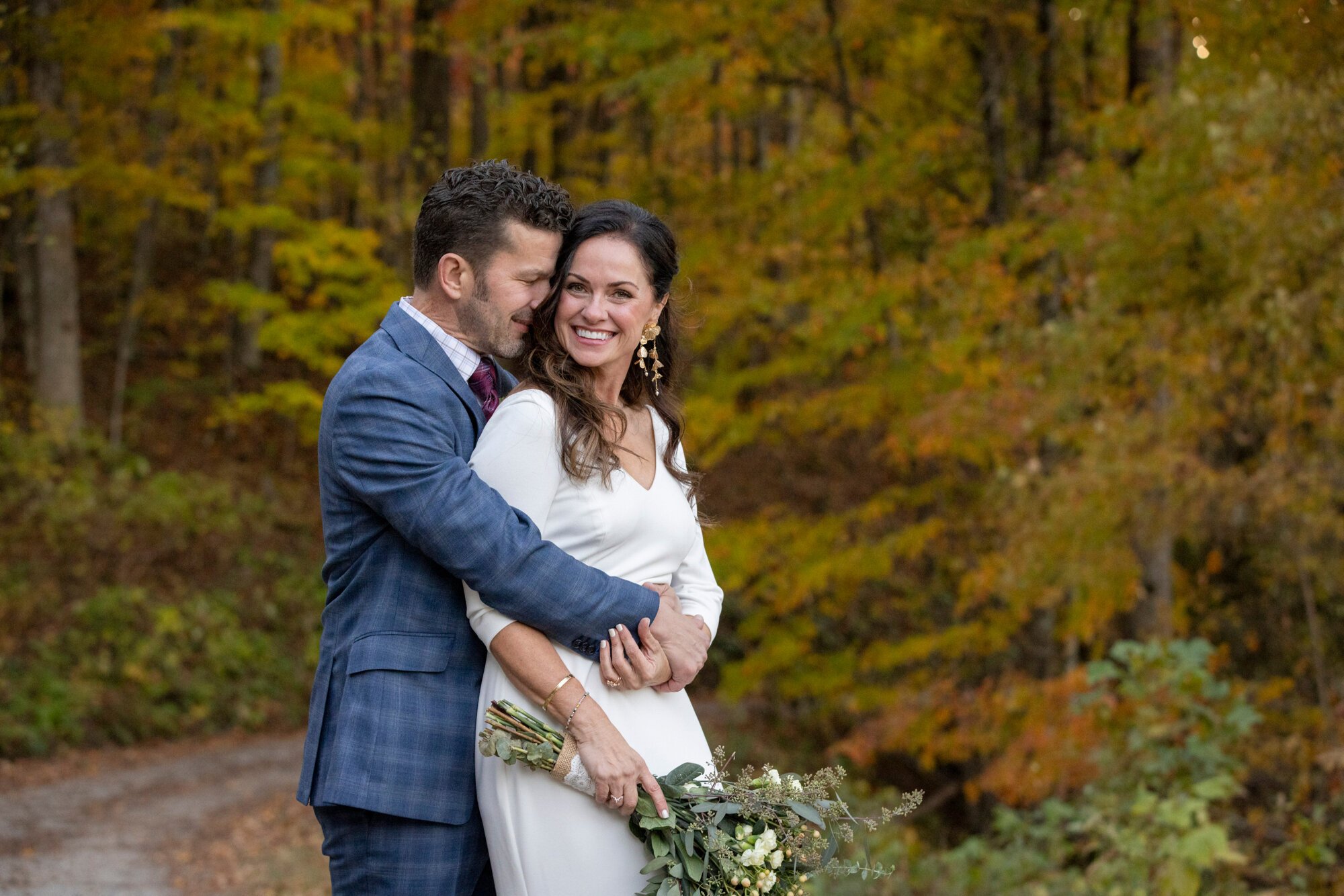 A happily smiling woman in a white dress holding a bouquet of flowers, embracing a man in a blue suit with curly brown hair, in outdoor autumn setting with colorful fall leaves in the background.
