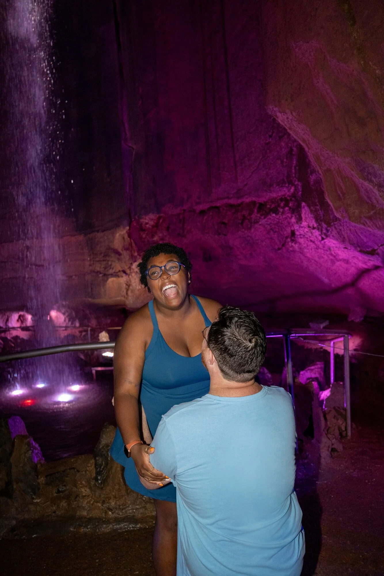 A woman in a blue top and glasses is smiling and talking to a man in a light blue shirt at an indoor waterfall with pink and purple lighting.