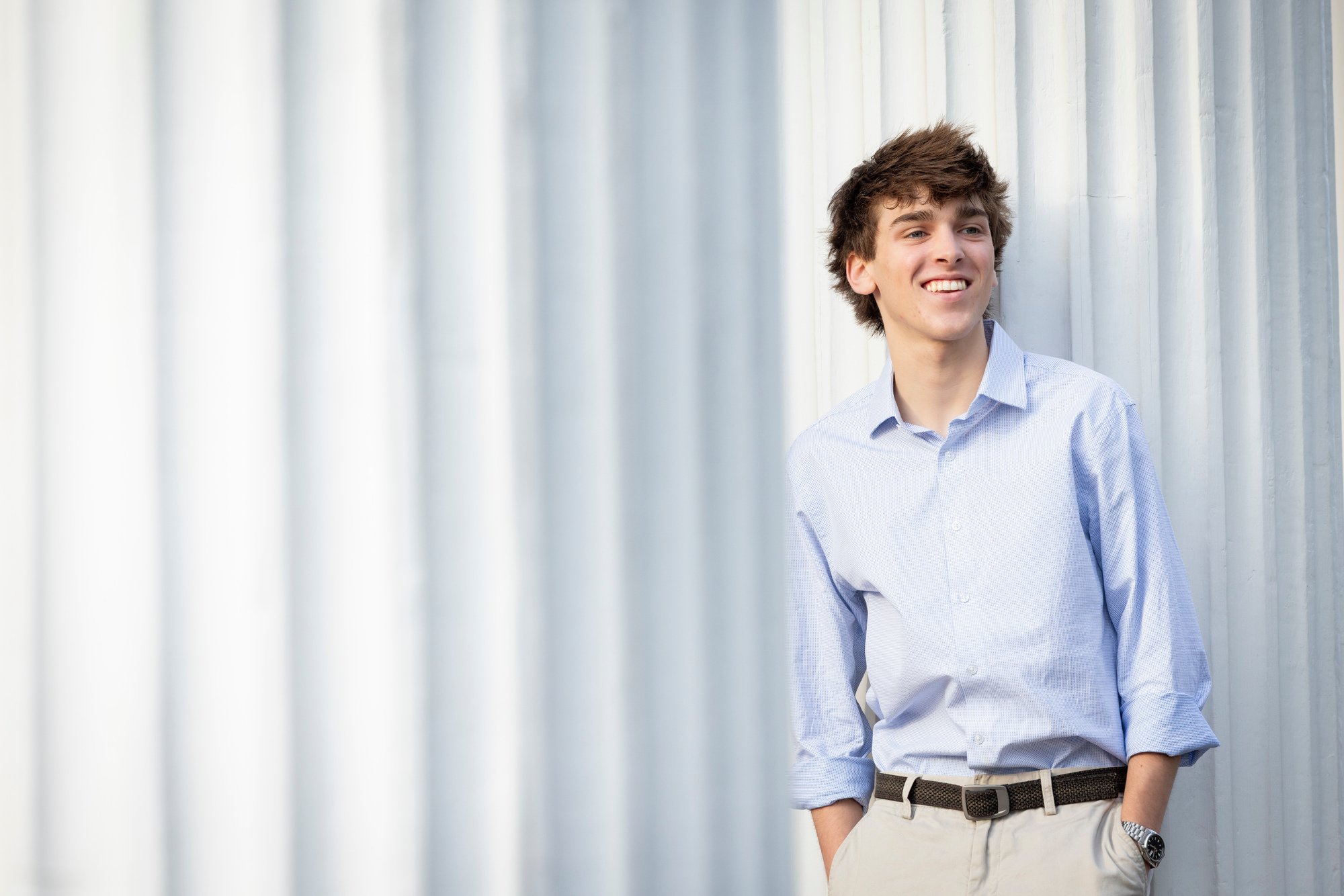 Young man with curly brown hair smiling, wearing a light blue button-up shirt and beige pants, leaning against a white corrugated metal wall.