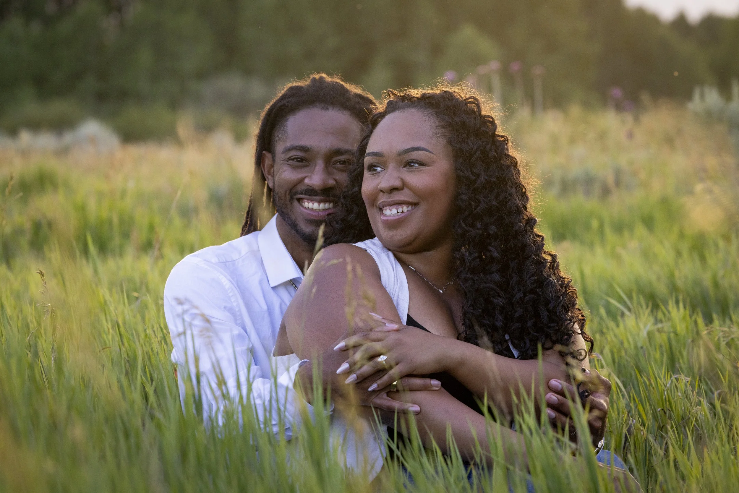 A smiling couple sitting close together in a grassy field during sunset, with the woman resting her head on the man's shoulder.