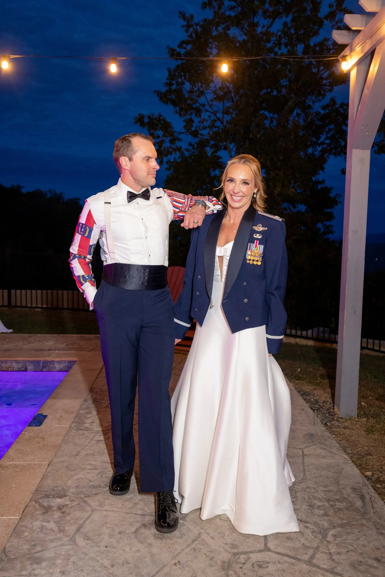 A man and woman dressed in formal and military attire, standing arm in arm outdoors at night under string lights, with trees and a dark evening sky in the background.