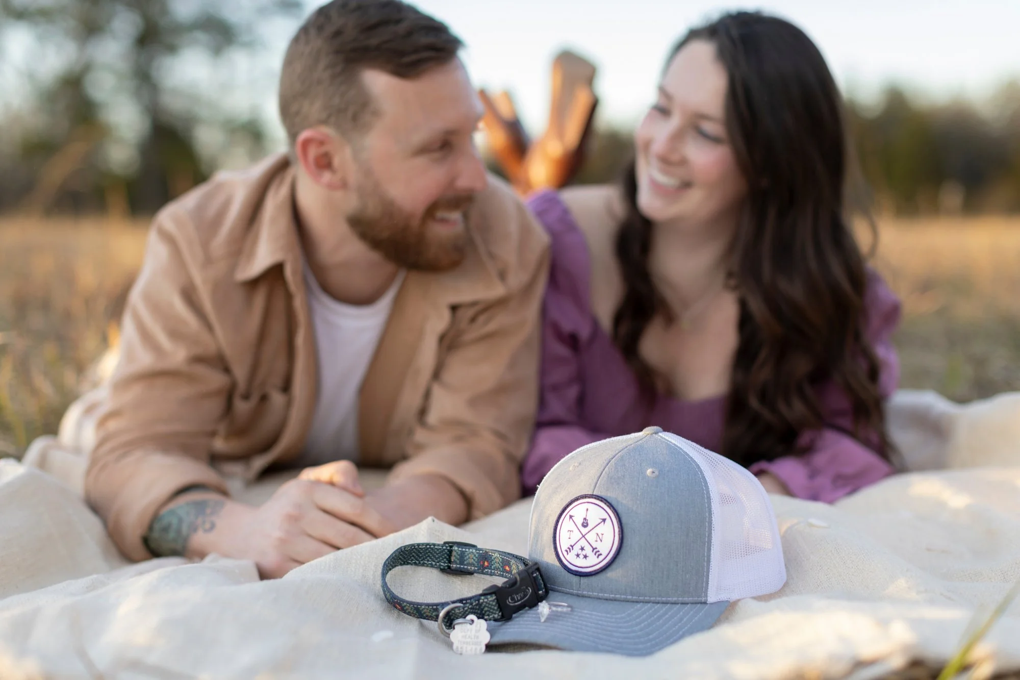 A smiling young man with a beard and a woman with dark hair sitting outdoors on the grass, having a conversation. A white cap with a purple logo and a buckle is in the foreground.