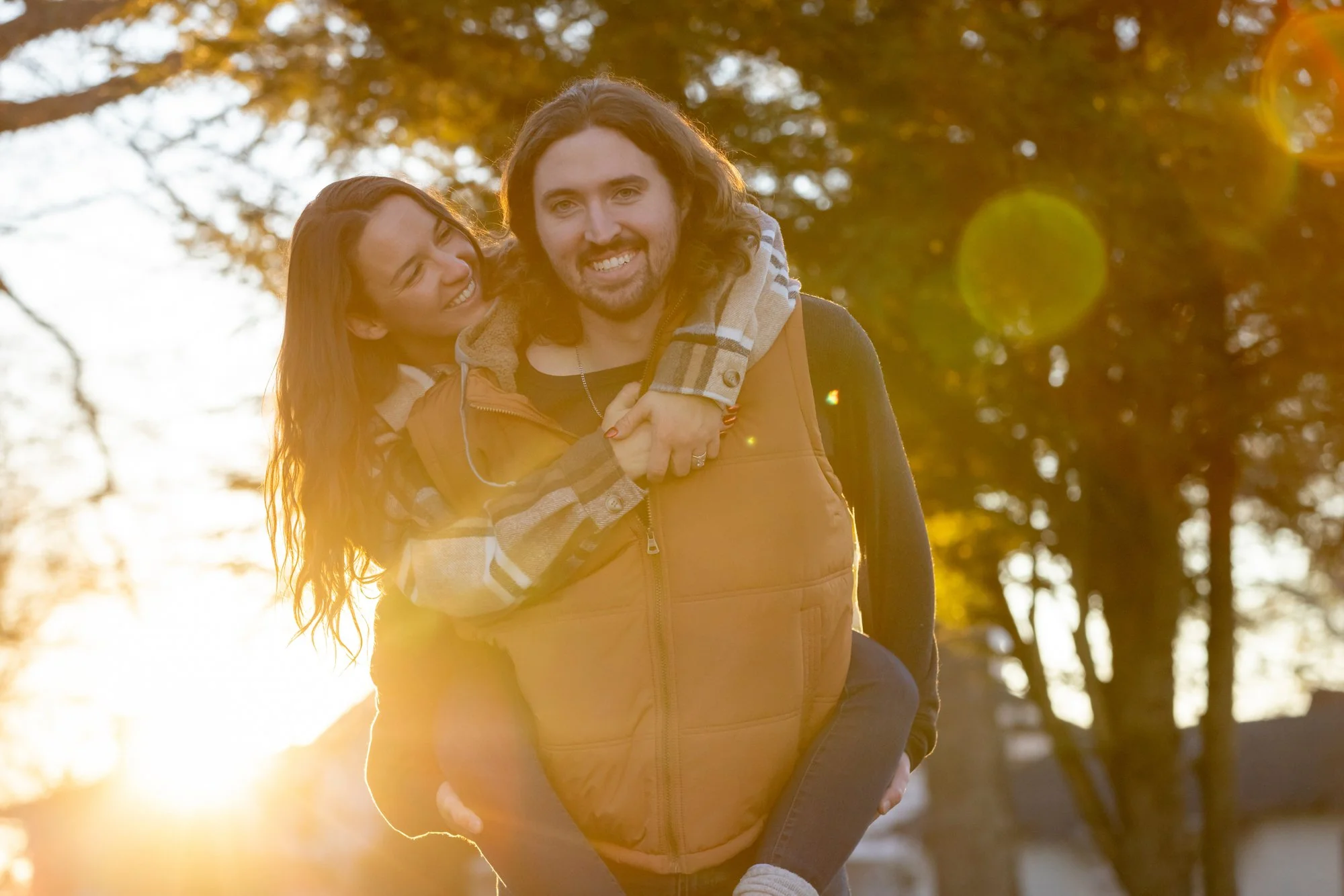 A couple enjoying a piggyback ride in a park during sunset, with fall foliage in the background.