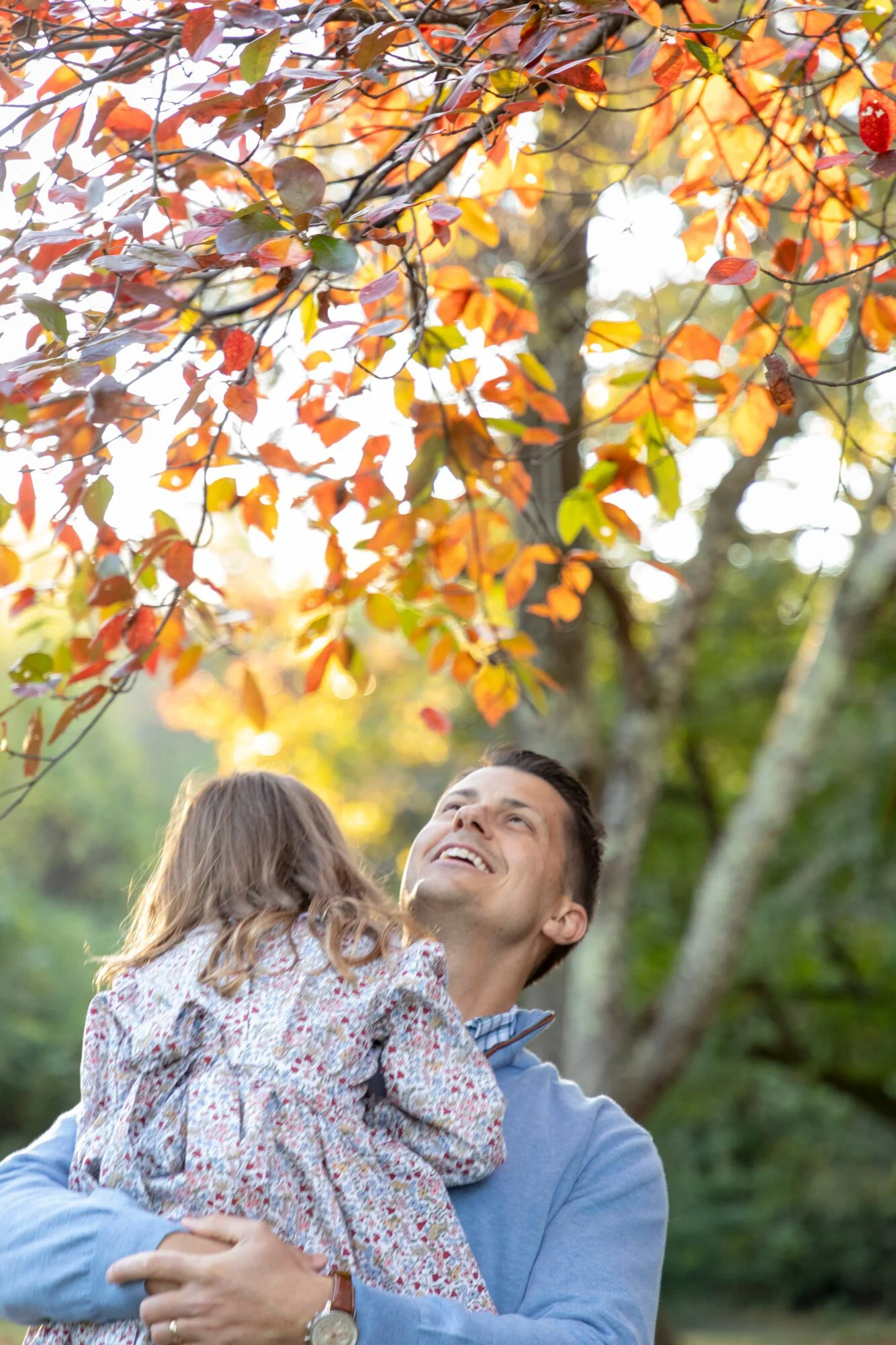 A man holding a young girl outside in a park with autumn leaves in the background.