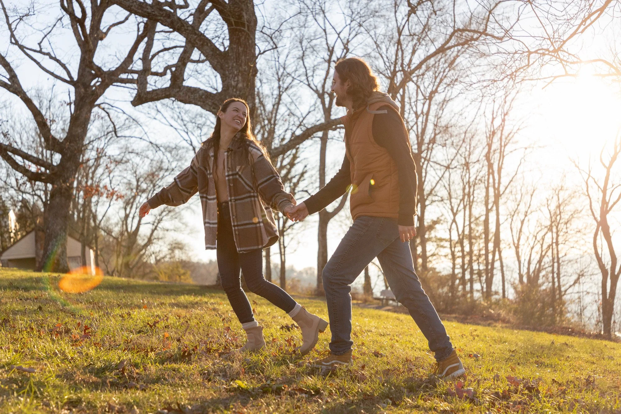 A couple holding hands and smiling while walking outdoors in a park during late afternoon or sunset. They are dressed casually, with the woman in a plaid jacket and the man in a brown vest. The scene features leafless trees and a grassy area.