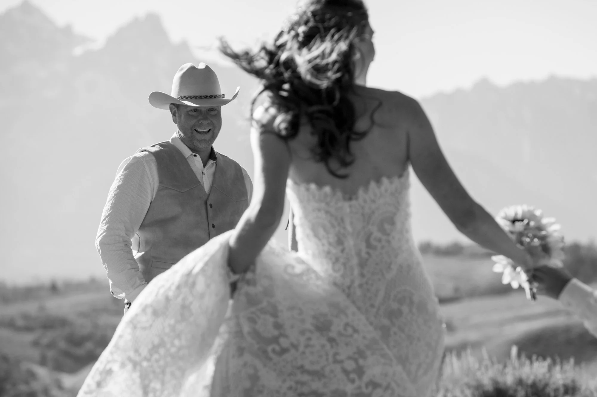 A man wearing a cowboy hat and vest smiling at a woman in a wedding dress on a sunny day outdoors.