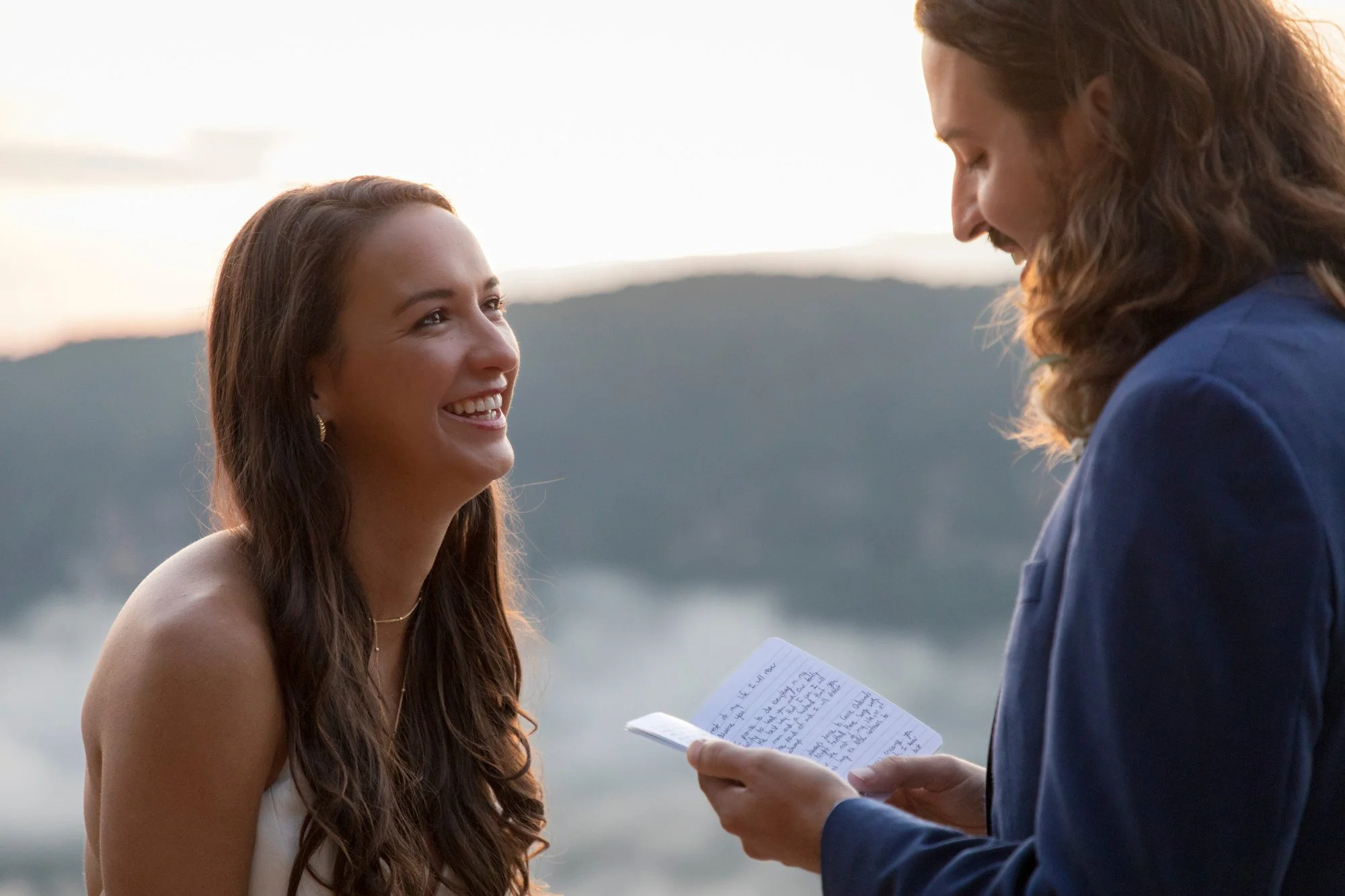 A woman with long brown hair smiling at a man with long wavy hair. The man is holding a small notebook with handwritten notes. They are outdoors during sunset with a mountain and lake in the background.