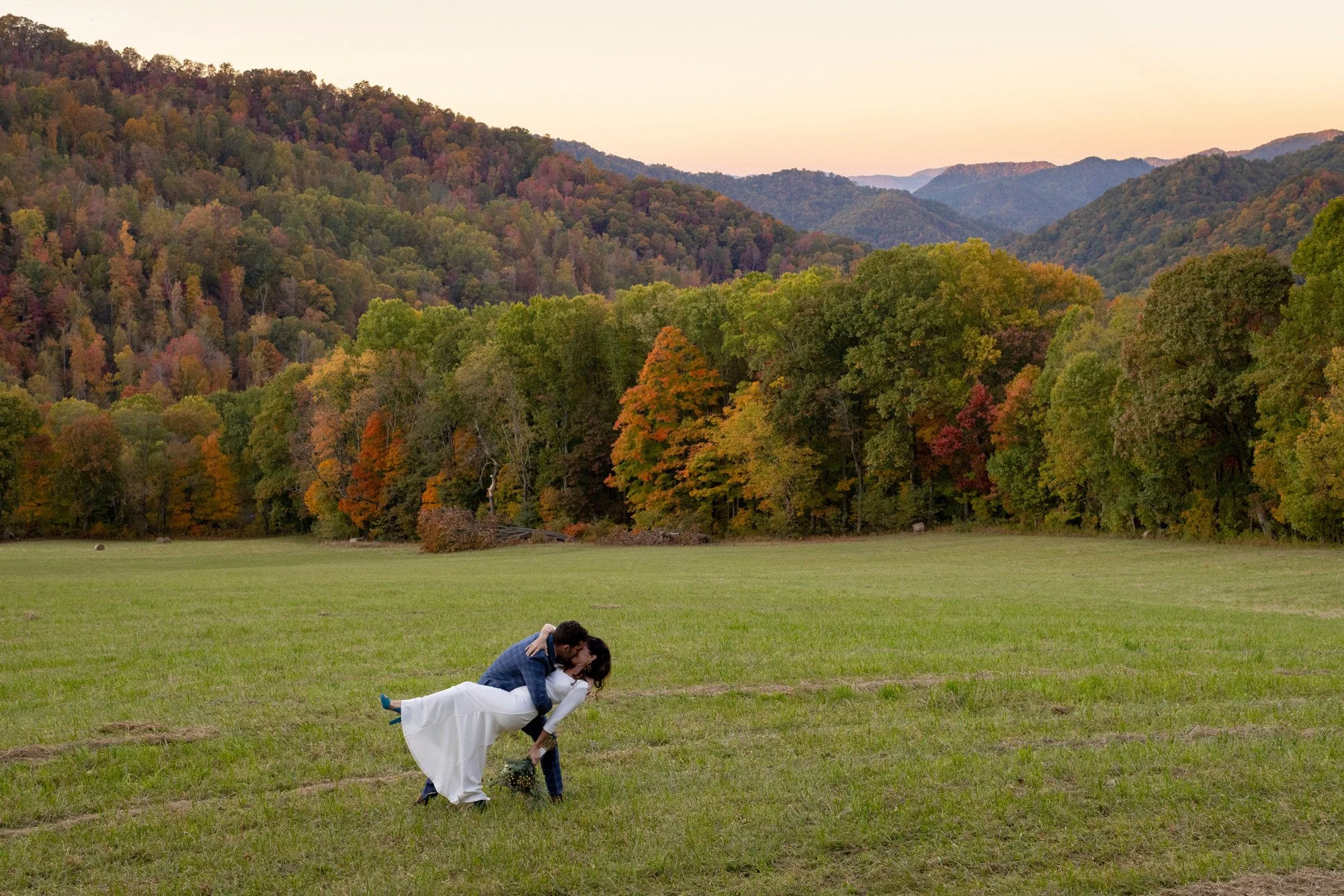 A couple is kissing in a field with mountains and colorful autumn trees in the background.