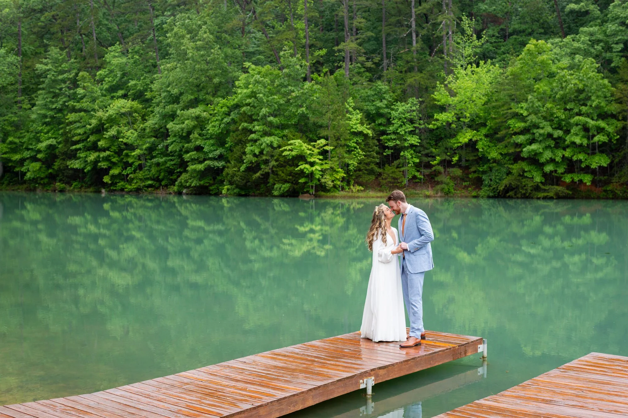 A bride and groom standing on a wooden dock, holding hands, with a lush green forest and a calm lake in the background.