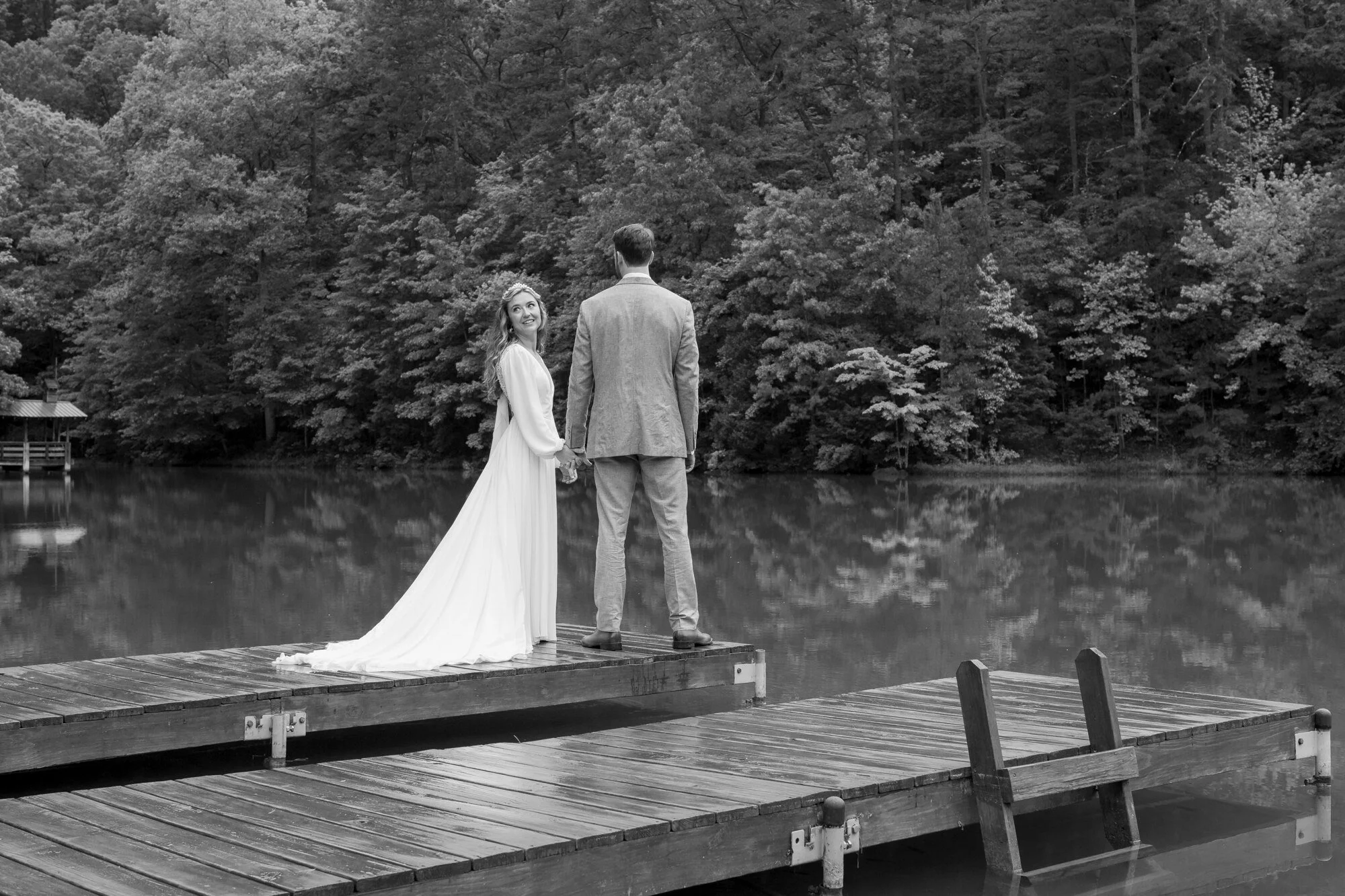 A bride and groom standing hand in hand on a wooden dock by a lake, with a dense forest in the background, in black and white.