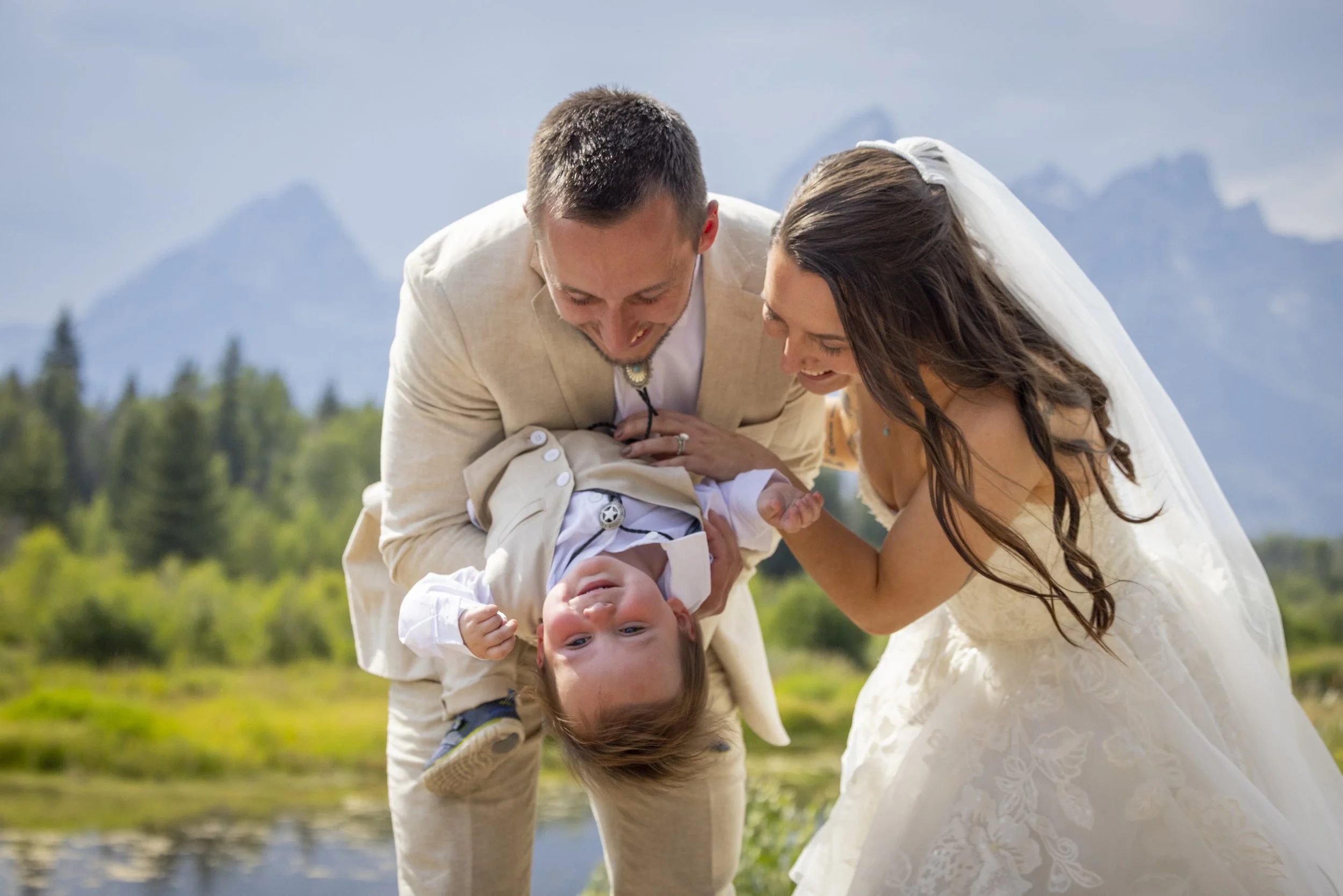 A couple in wedding attire bending over outdoors, holding their young child upside down, with mountains and trees in the background.