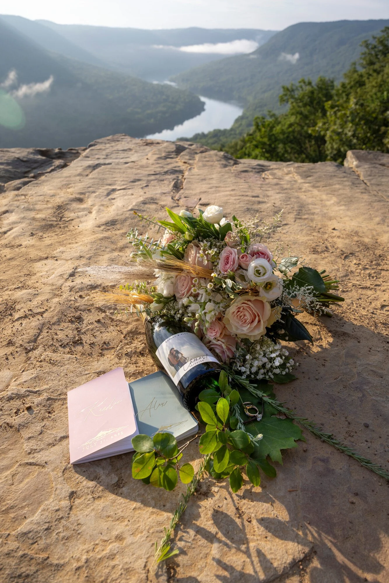 A bouquet of pink and white roses with green leaves and small white flowers rests on a rocky surface, with a scenic view of a river and mountains in the background under a cloudy sky.