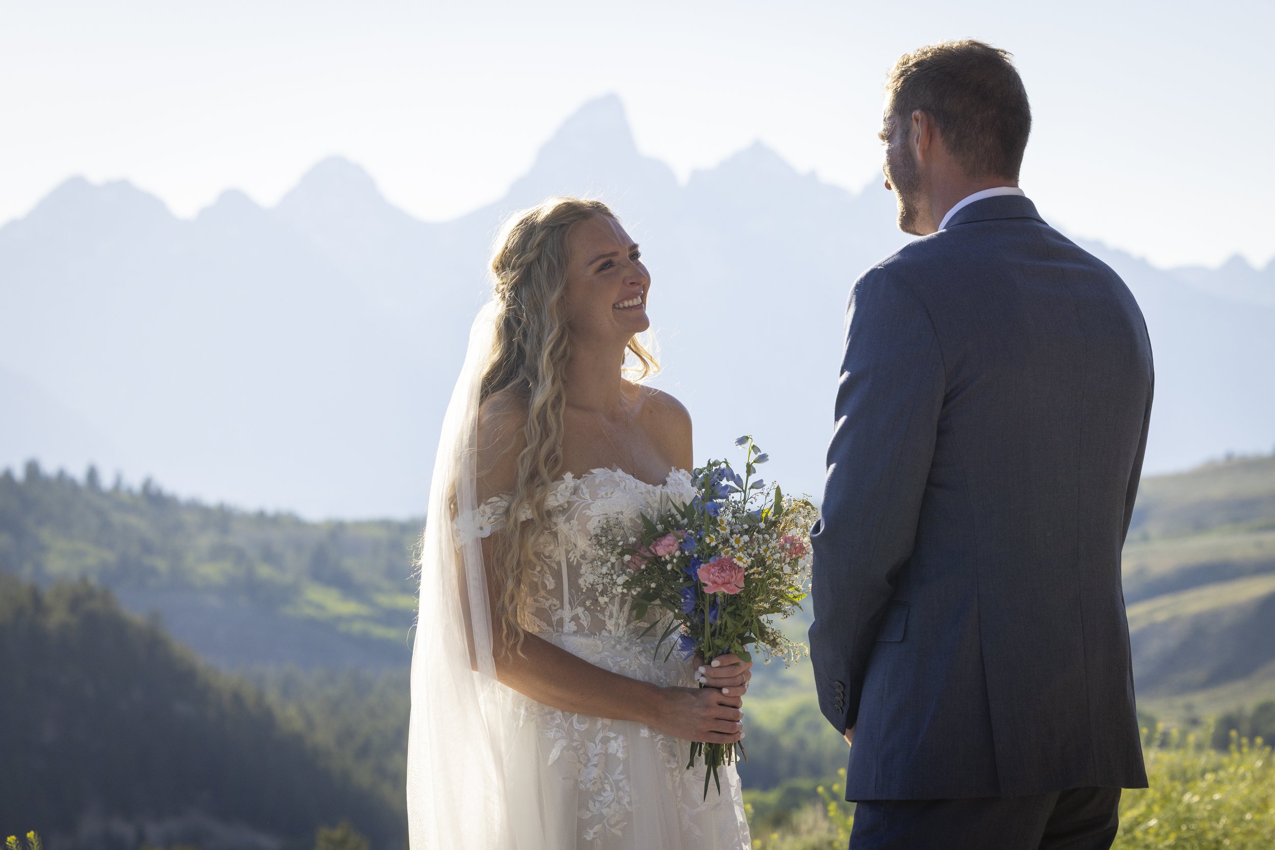 A bride in a white lace wedding dress holding a bouquet of colorful flowers, standing and smiling at a groom in a blue suit outdoors with mountains and sky in the background.