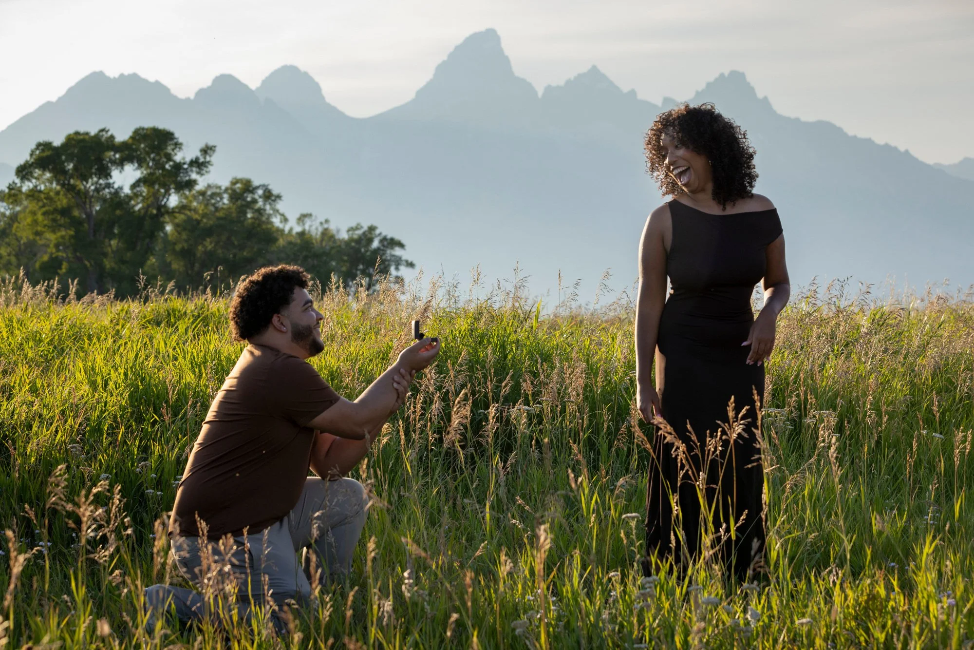 A man kneeling in a grassy field, taking a photo of a woman standing in front of him, with a backdrop of trees and mountains.