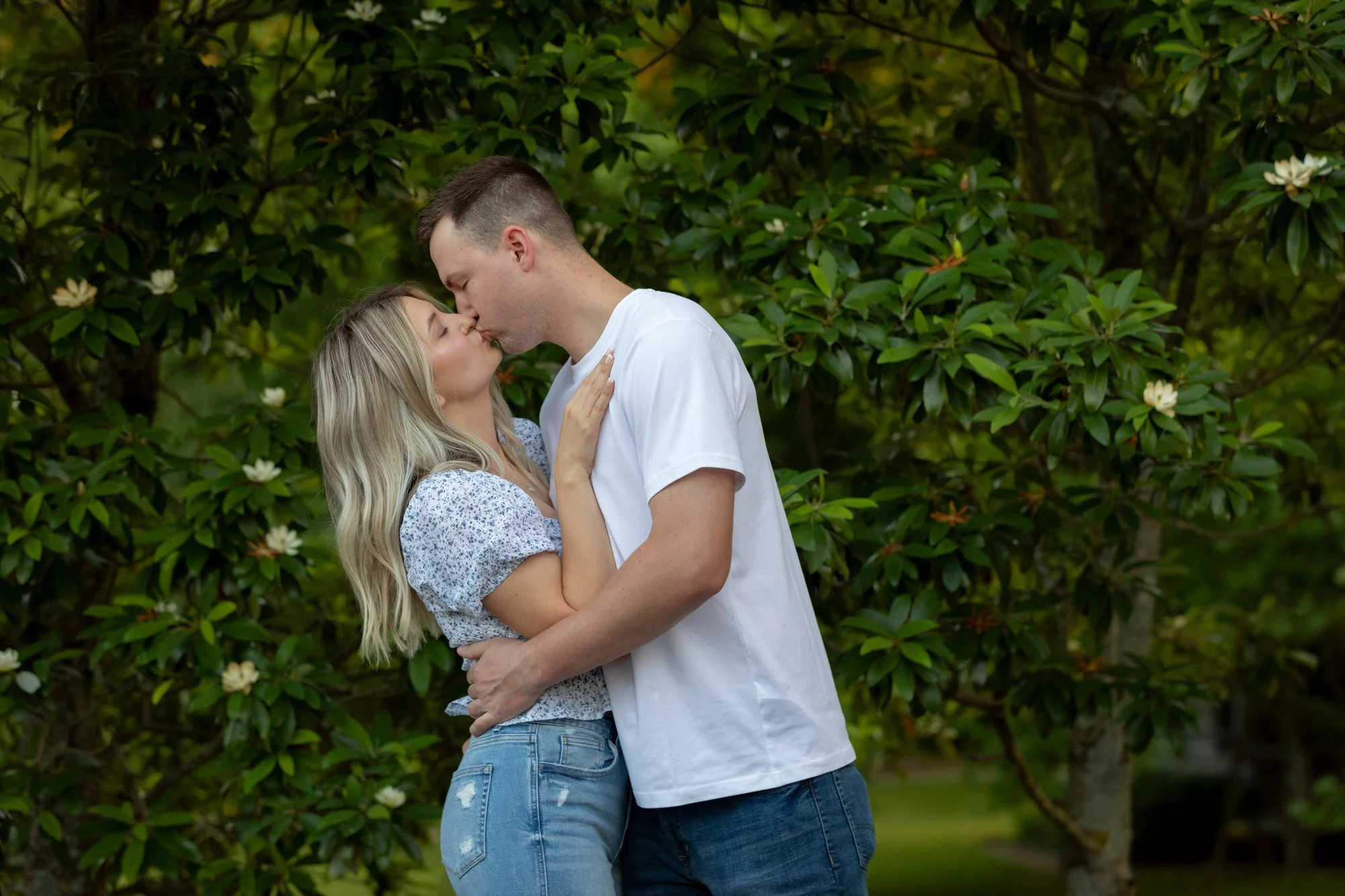 A couple kissing outdoors in front of green leafy bushes with white flowers, the woman has blonde hair and wears a patterned top, and the man has short brown hair and wears a white t-shirt.