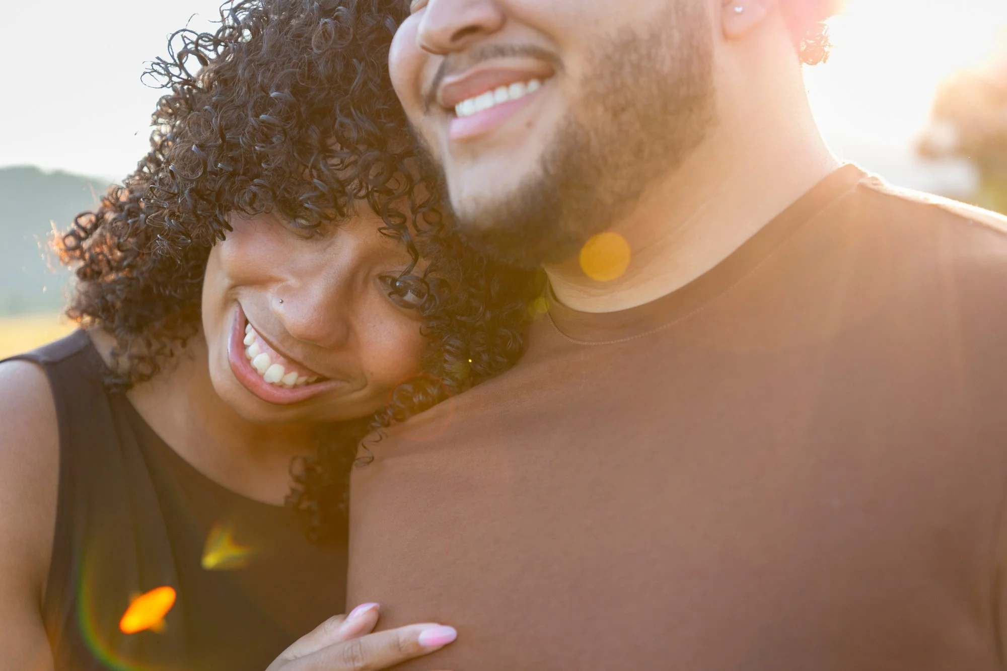 A close-up of two people hugging outdoors in a sunny setting, with one person smiling and the other's face partially visible, showing curly hair and a nose piercing.