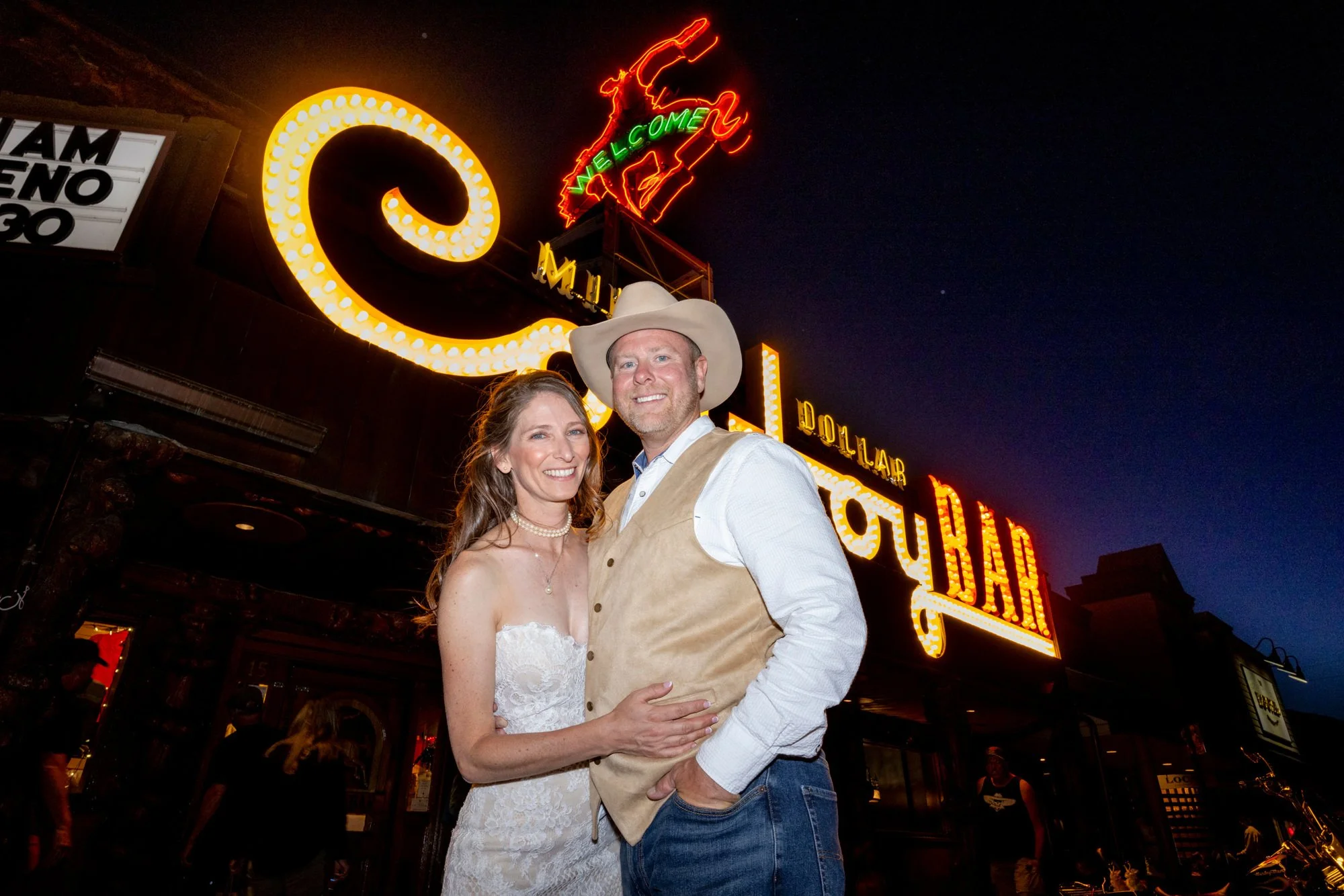 A happy couple posing together outside at night with neon signs in the background, one sign reads 'Welcome' and the other 'Honky Tonk.' The woman is wearing a white lace dress and the man is wearing a cowboy hat, beige vest, and white shirt.