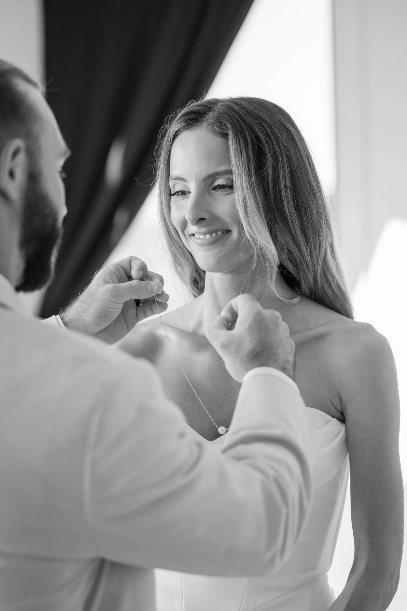 A woman smiling as a man helps her with a necklace, in a black-and-white photo.