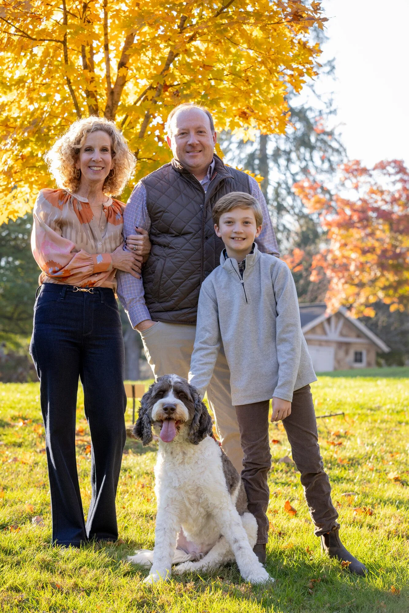 A family of three, a woman, a man, and a boy, standing outdoors in a park with autumn trees in the background. The woman is smiling and holding the man's arm, the man is smiling with hands in pockets, and the boy is smiling and standing beside a larg