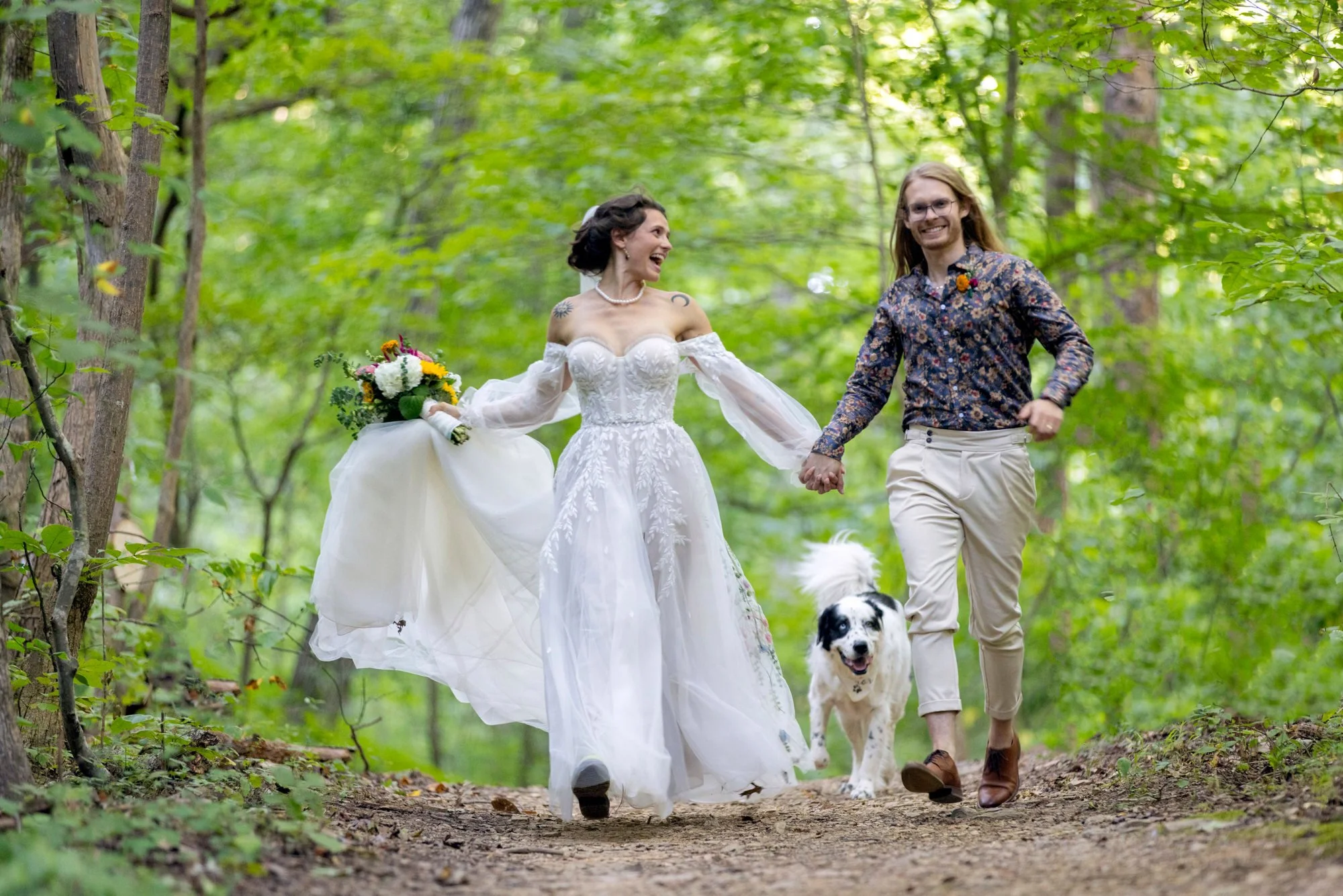 A couple, dressed in wedding attire, walking in a lush, green forest holding hands. The bride wears a white wedding dress and the groom wears a patterned shirt with beige pants. They are accompanied by a dog walking alongside them, and both are smili