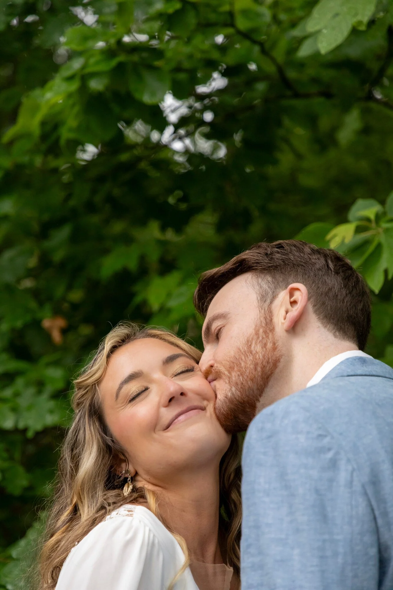 A couple sharing a kiss outdoors, with the woman smiling and eyes closed, surrounded by green foliage.