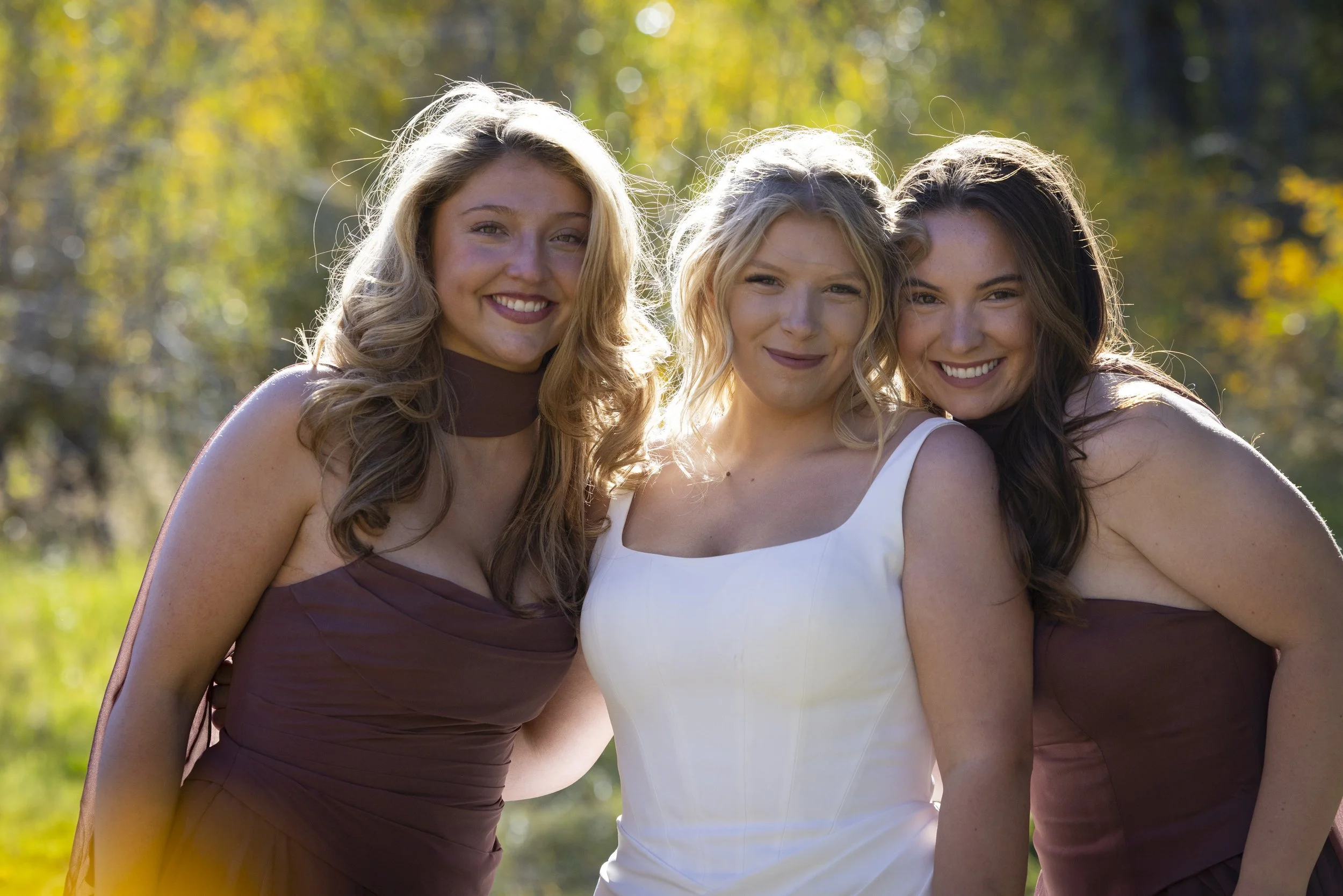 Three women smiling and standing close together outdoors in a sunny, autumn setting.