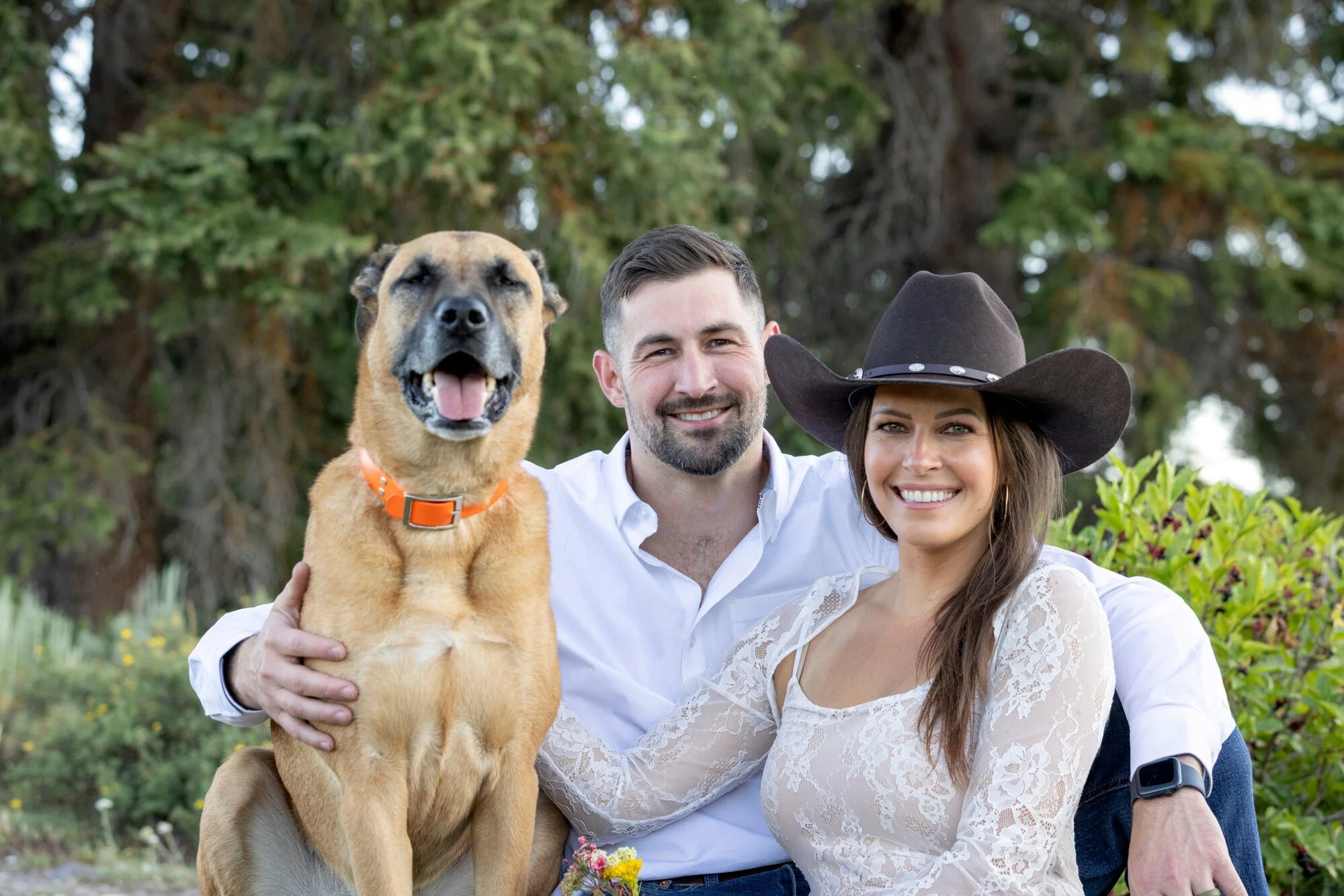 A smiling couple, a woman wearing a large black cowboy hat and a man with short dark hair and a beard, pose outdoors with a large happy dog with a tan coat and black face markings, in front of a green leafy background.