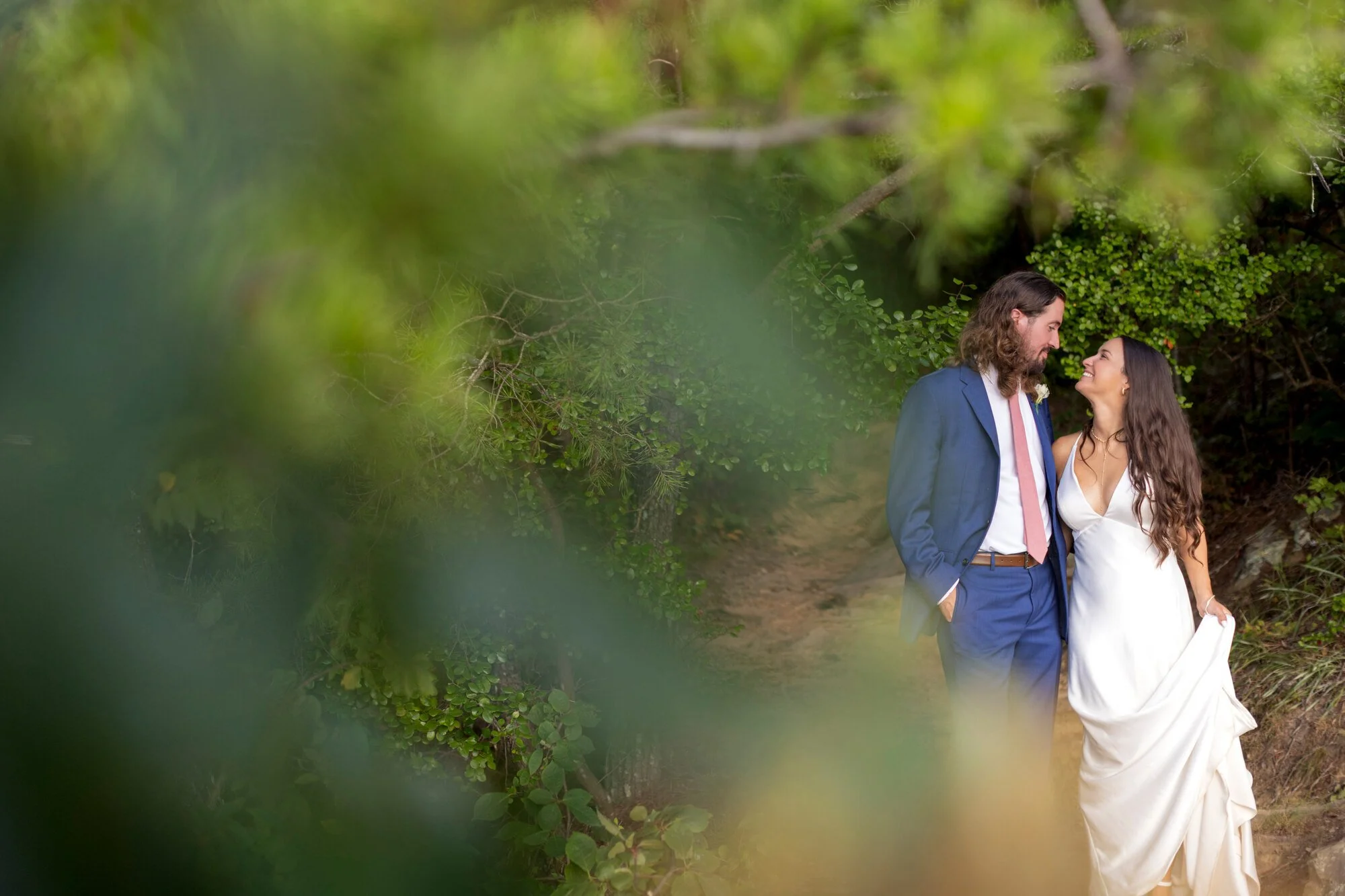 A couple in wedding attire standing close together outdoors amidst green foliage, smiling at each other.
