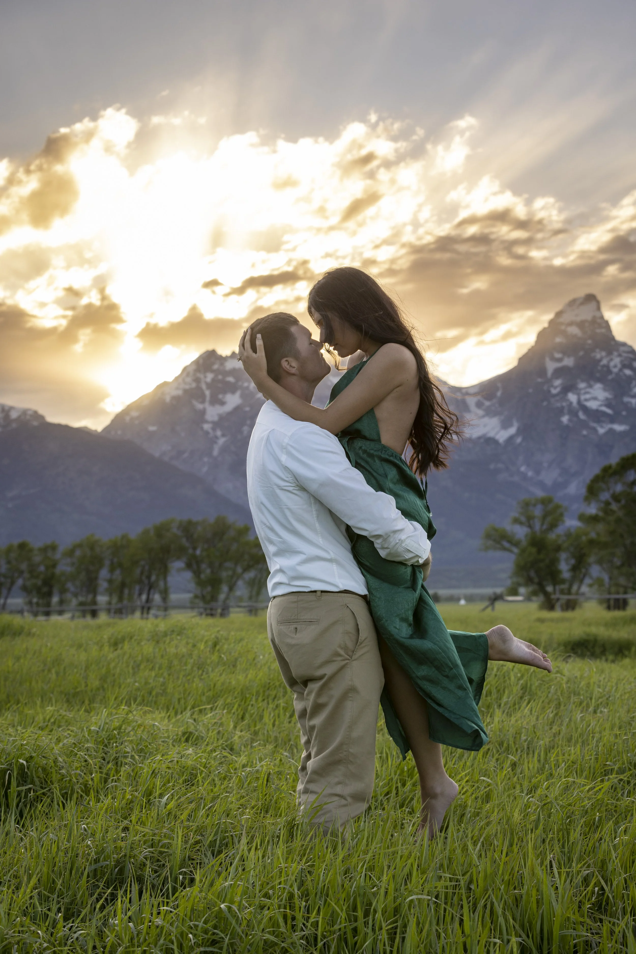 A man lifting a woman in a grassy field during sunset with mountains in the background.