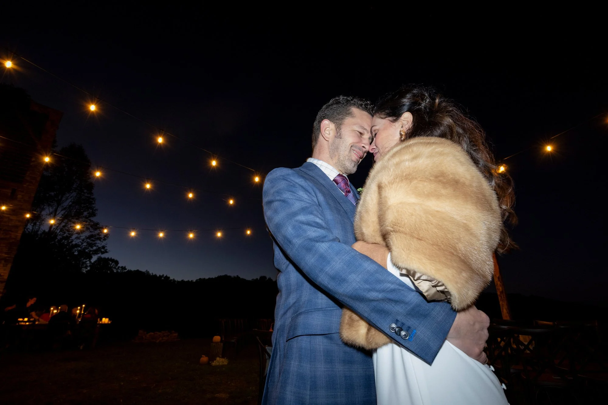 A man and woman embrace closely with their foreheads touching at night, under string lights, outside. The man is dressed in a blue suit with a dress shirt and tie. The woman is wearing a white dress and a fur shawl. The background shows dark sky, tre