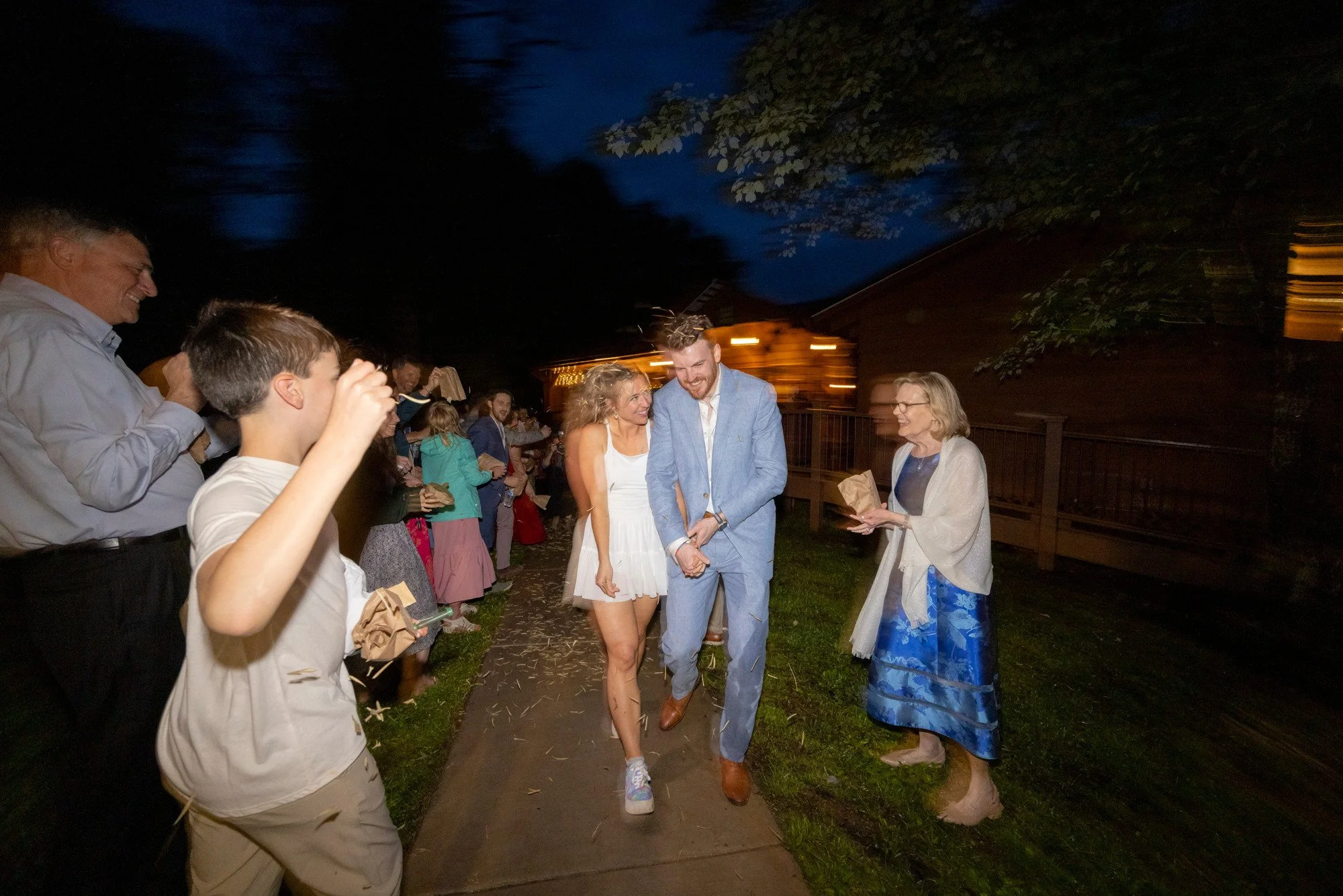 A group of people at an outdoor evening event, with a couple holding hands at the center, smiling and surrounded by others. The background shows a dark sky and trees with some lights and a small building.