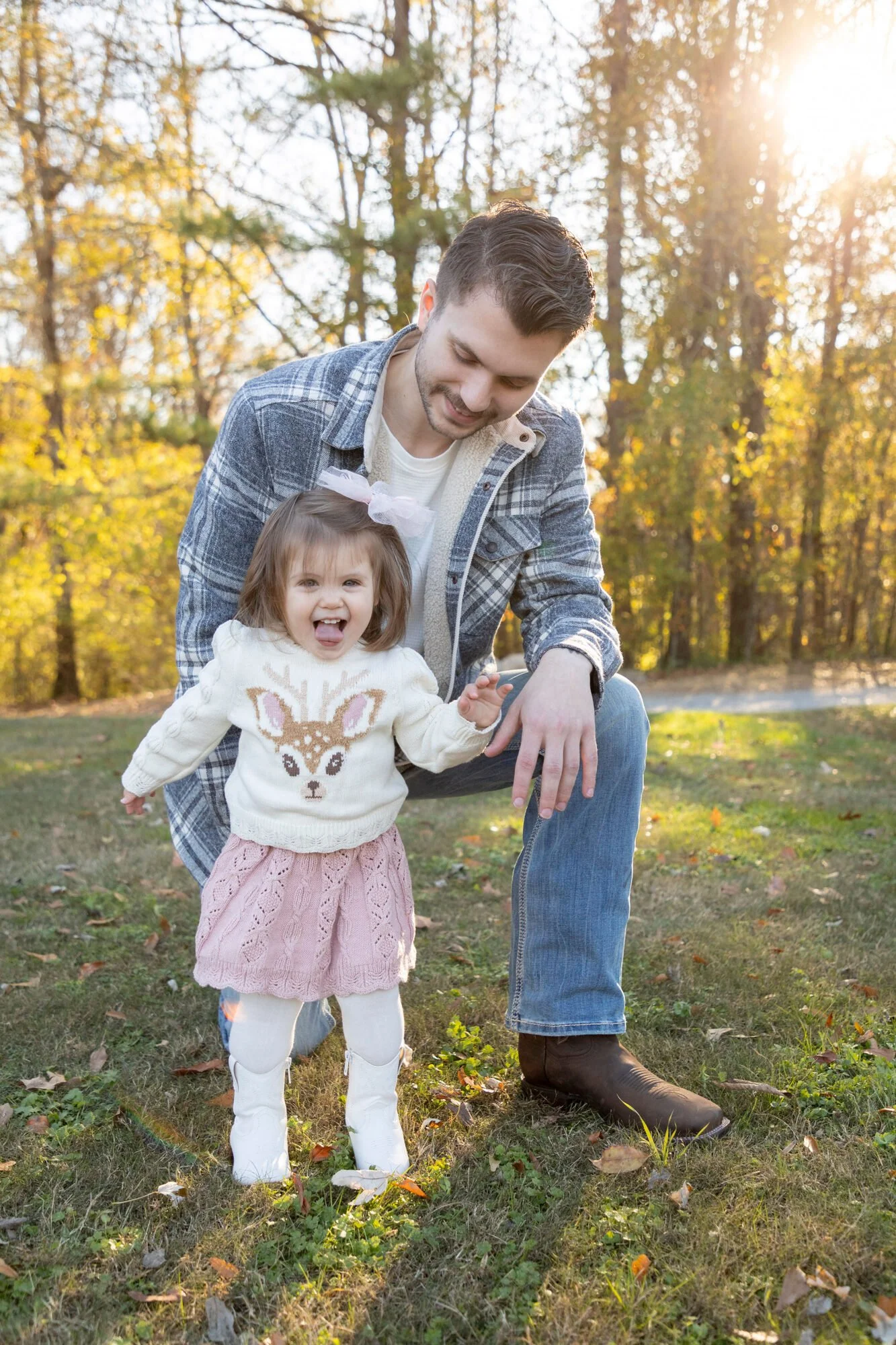 A man and a young girl are outdoors in a park during fall, with trees full of yellow leaves and sunlight filtering through. The girl is smiling and sticking her tongue out, wearing a cream sweater with a reindeer design, a pink skirt, and white tight