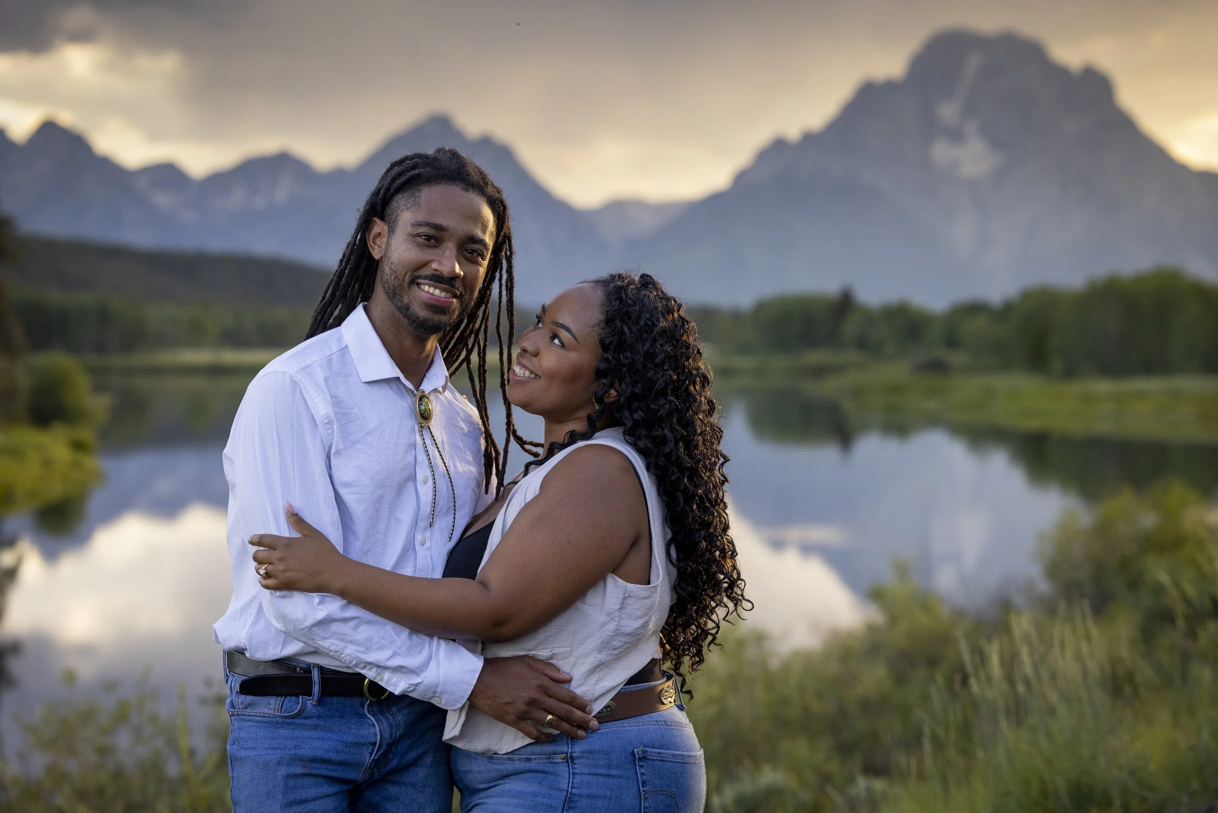A couple embraces outdoors with mountains and a lake in the background during sunset, smiling at each other.