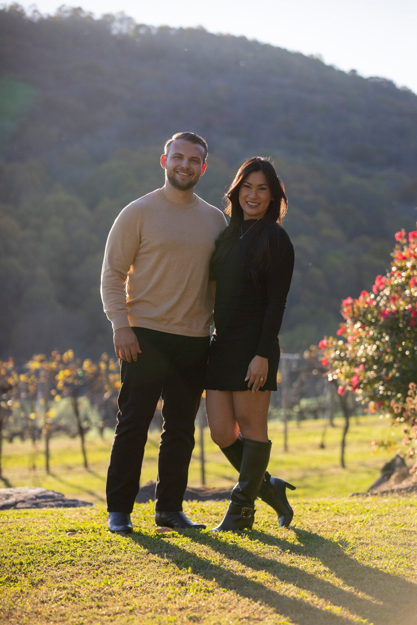 A smiling man and woman stand together outdoors in a park during daylight, with a hill and flowering bushes in the background.