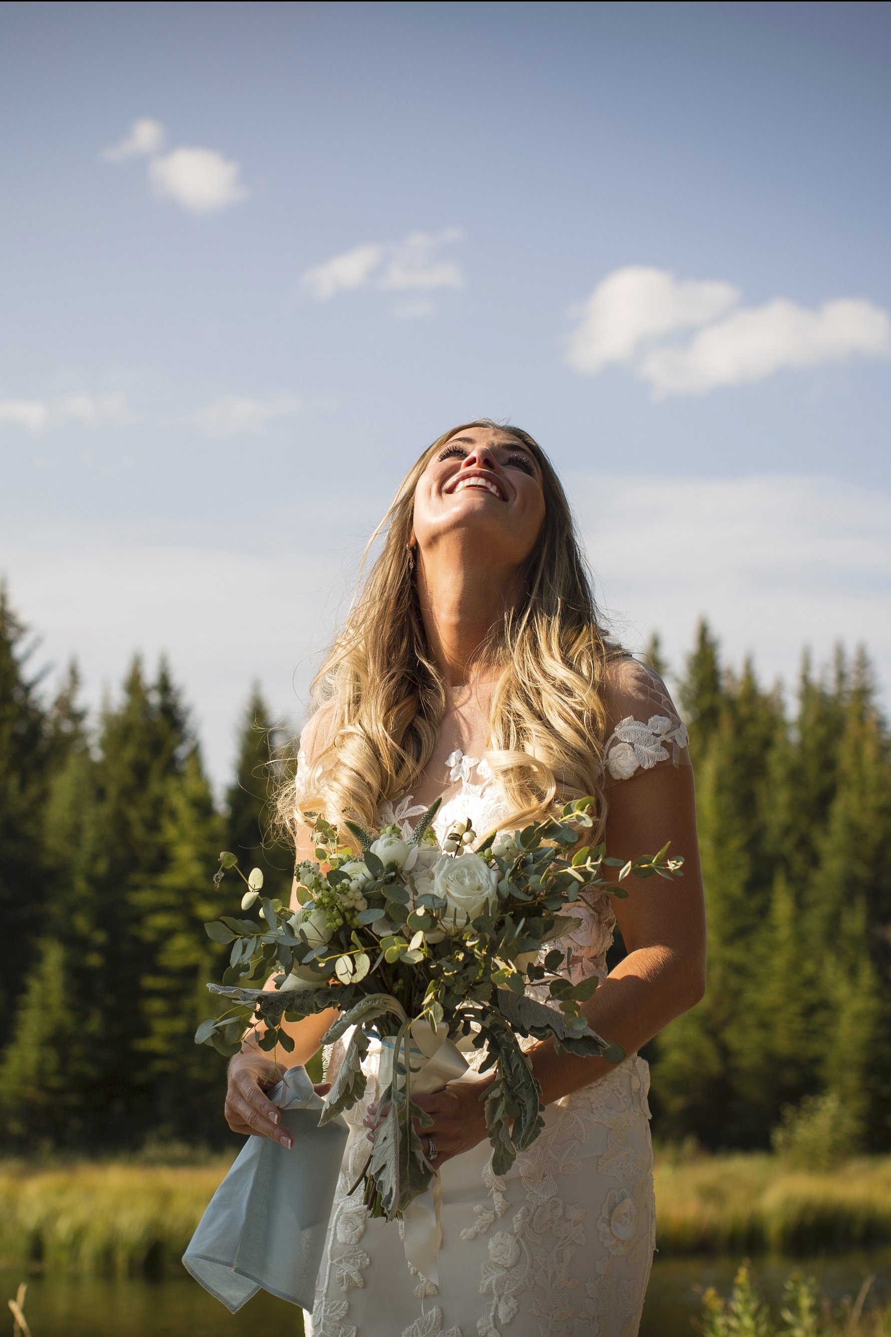 A smiling woman in a wedding dress holding a bouquet of flowers outdoors in a natural setting with trees and a blue sky with clouds in the background.